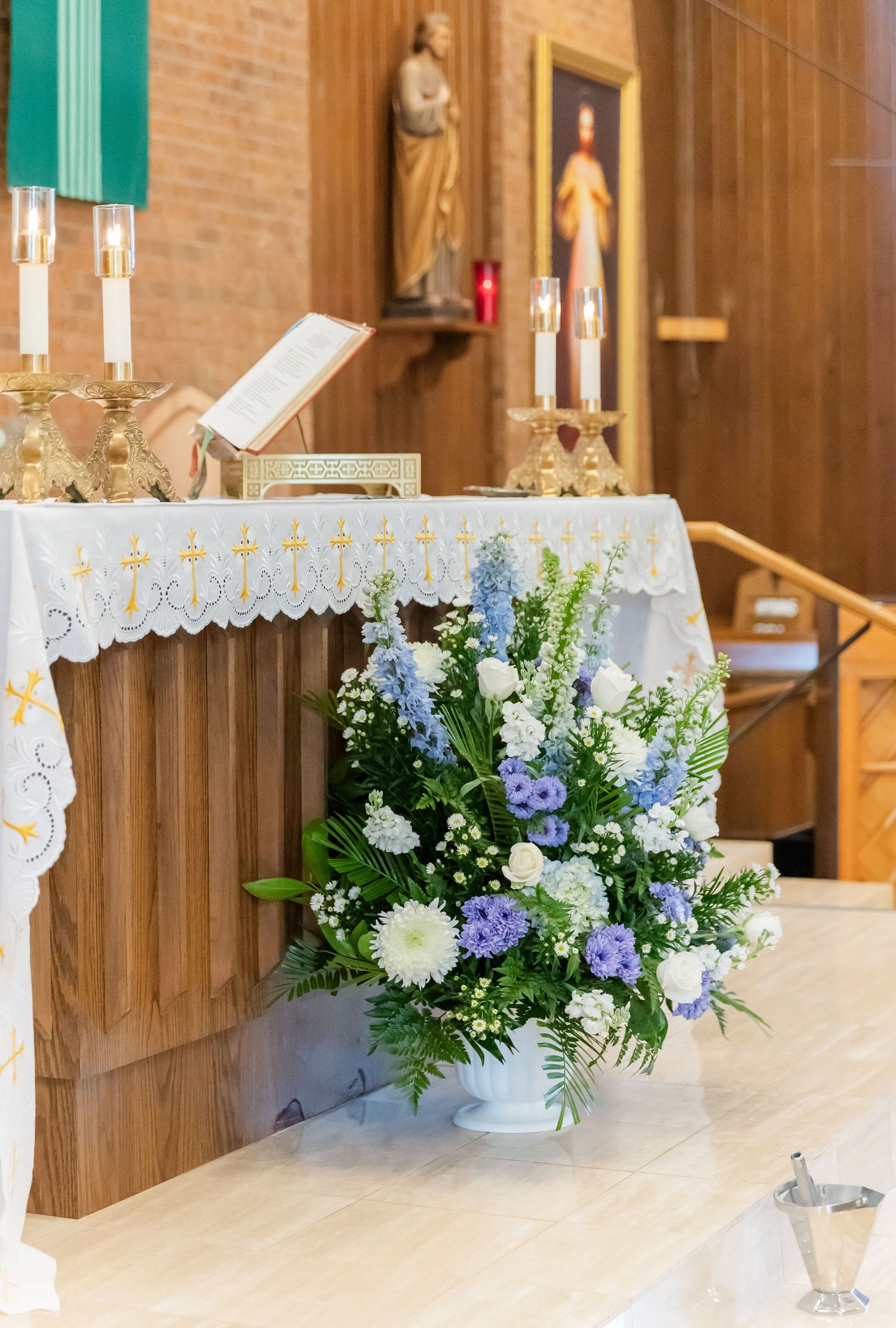 Church altar decorated with a white lace cloth and blue and white floral arrangement with candles and religious icons.