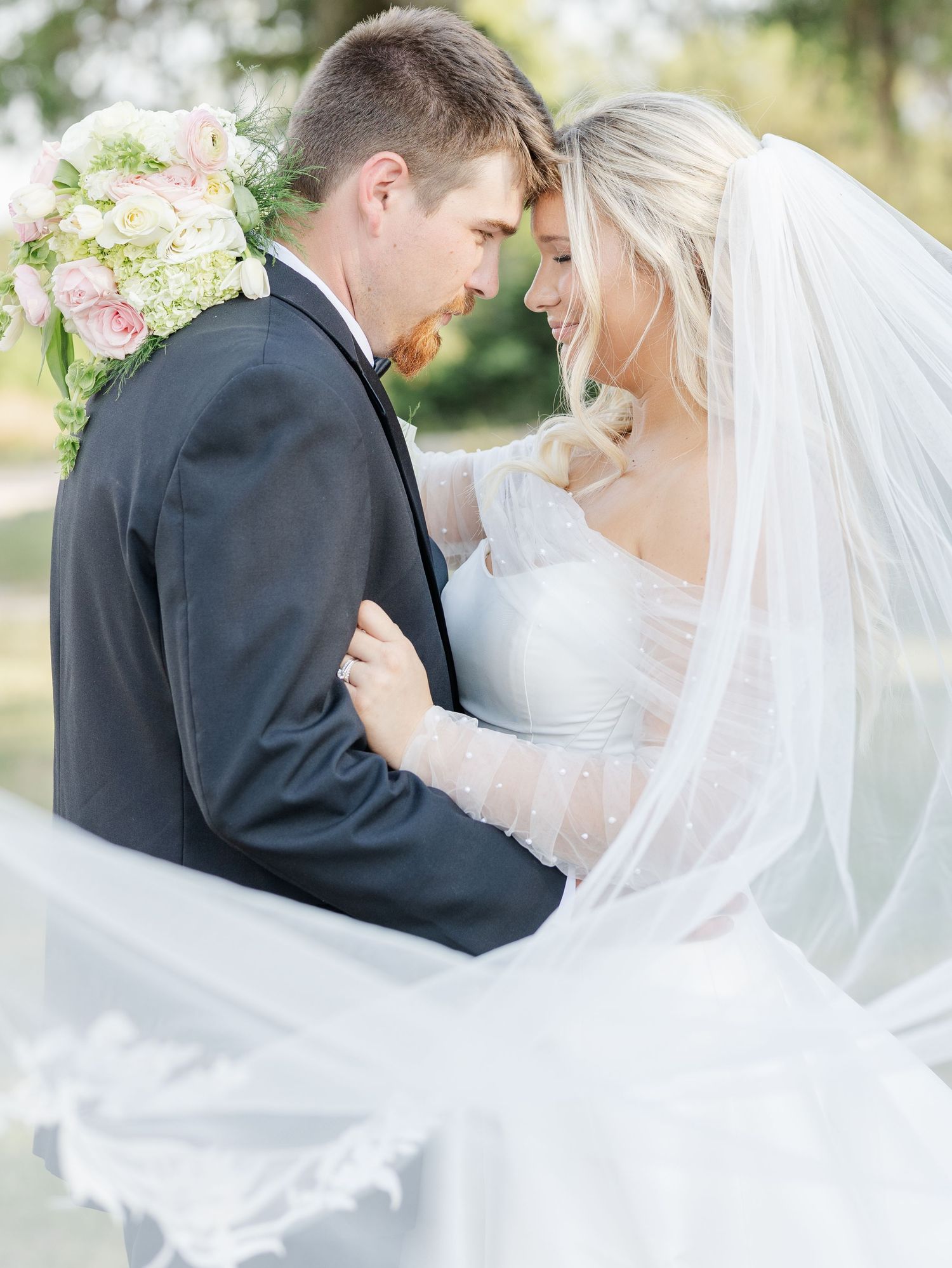 A bride and groom share a romantic moment, her veil flowing, as she smiles in a white gown with a floral bouquet nearby.