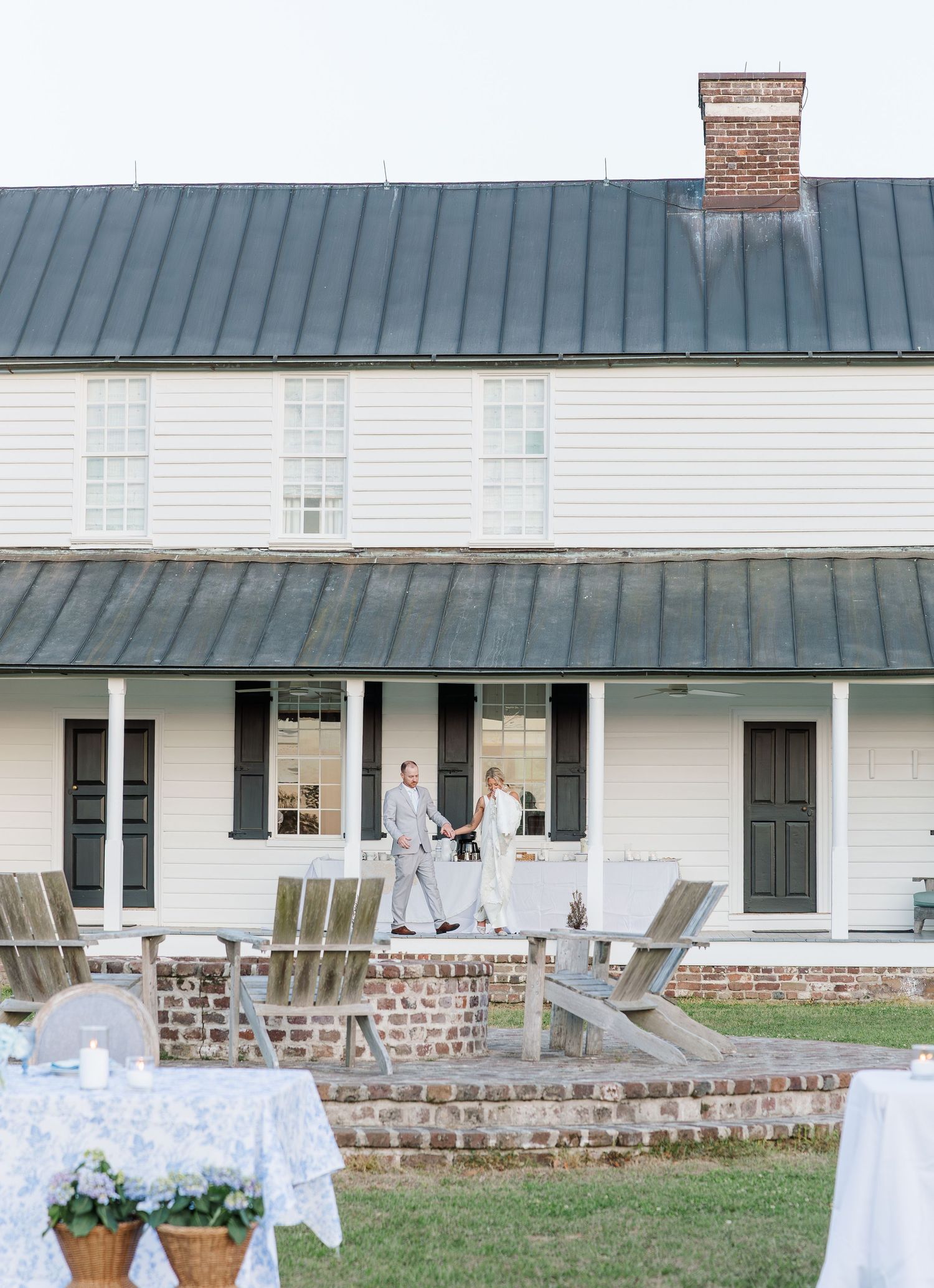 Bride and groom hold hands on porch of white historic farmhouse wedding venue at dusk.