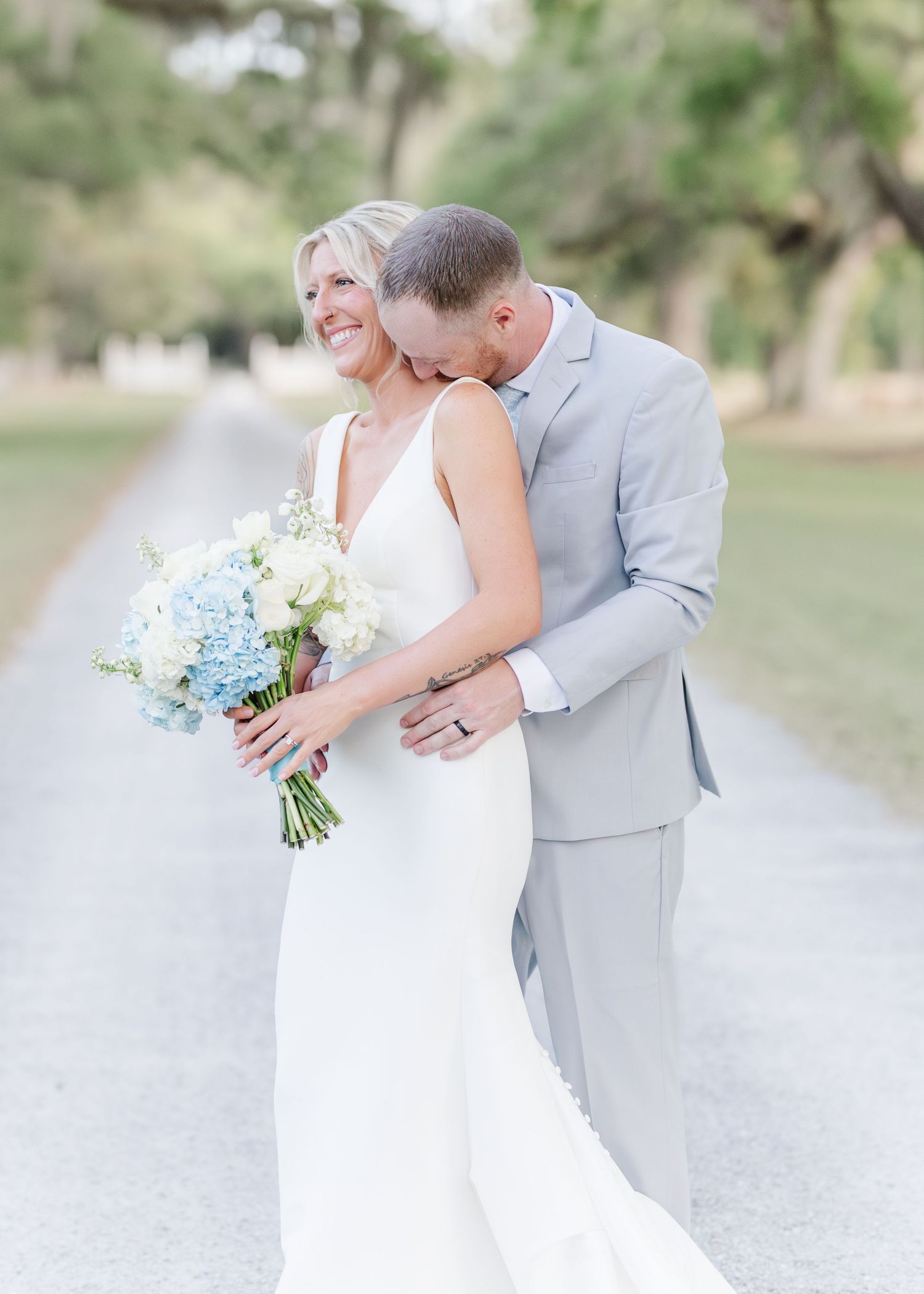 Bride holding blue hydrangea bouquet while groom kisses her cheek outdoors.