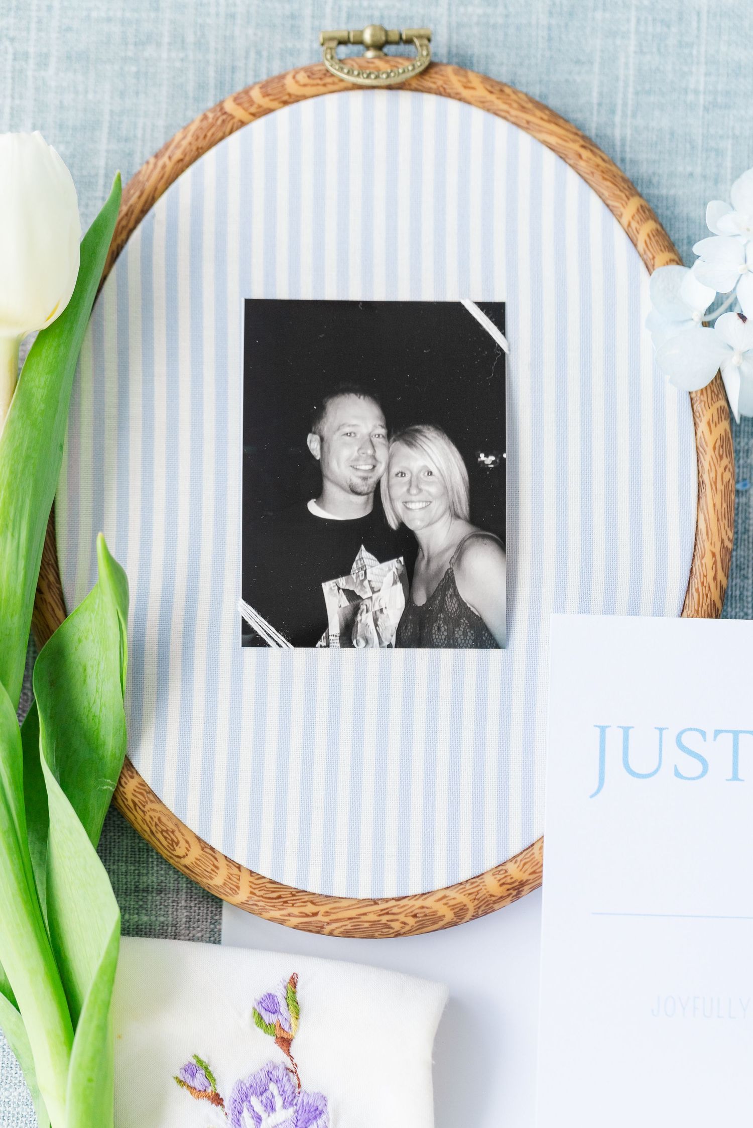 Oval embroidery hoop frame with black and white couple photo on blue striped fabric beside white tulip and wedding stationery.