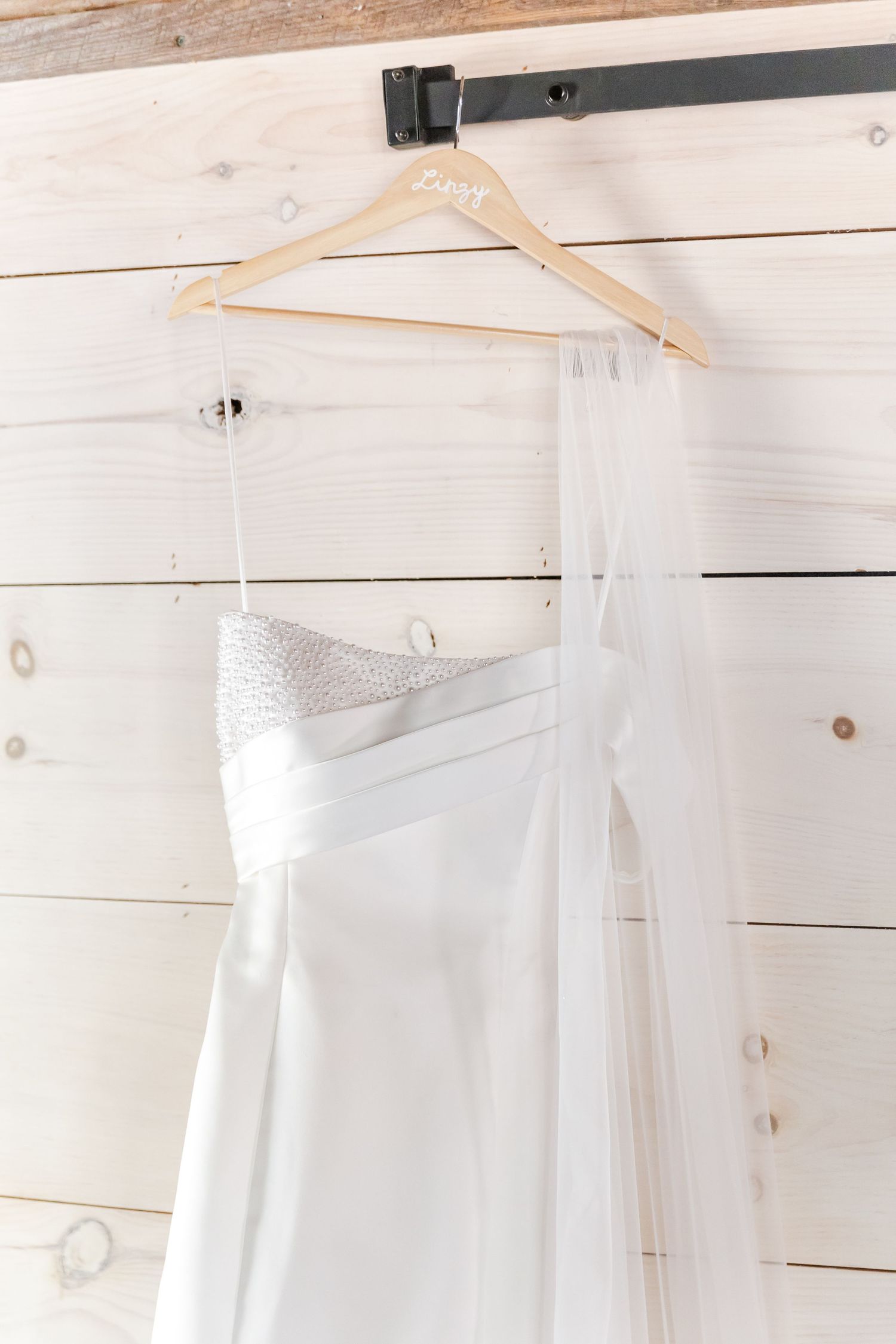 Elegant strapless white wedding gown with beaded bodice and veil hanging on personalized wooden hanger.