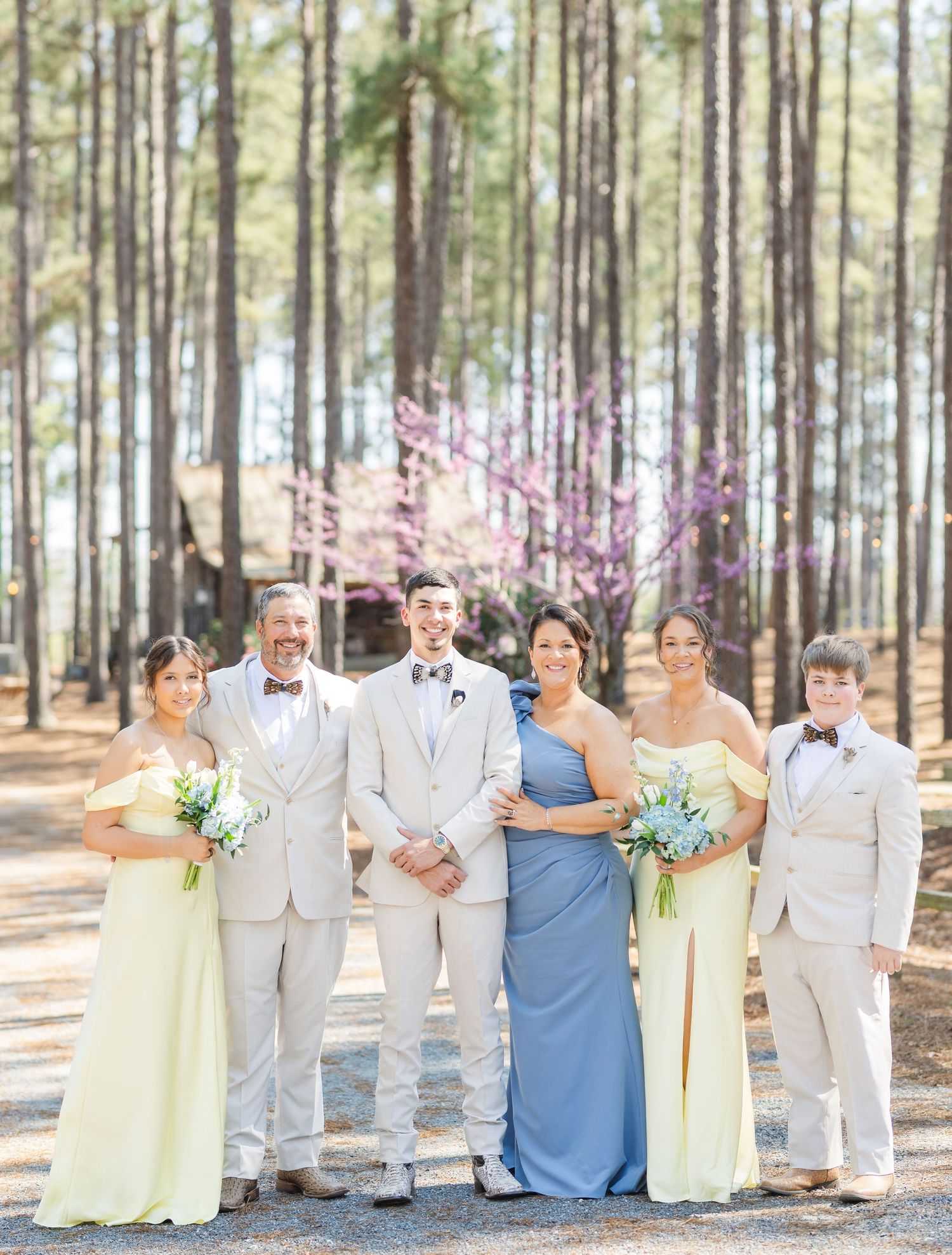 Groom with wedding party in yellow and blue bridesmaid dresses among pine trees with redbud blooms.
