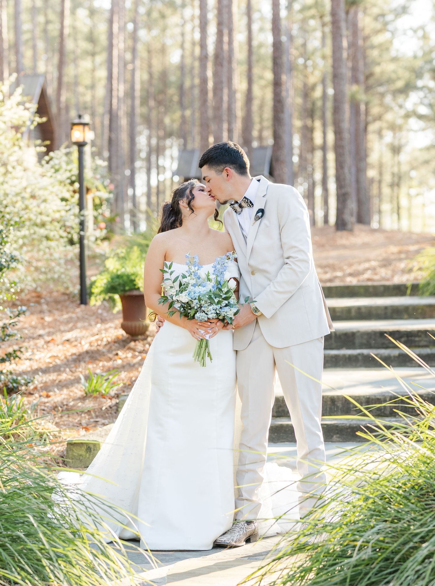 Bride and groom share a romantic kiss on stone steps surrounded by tall pine trees and lush greenery.