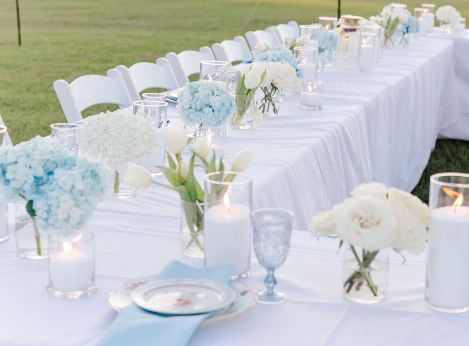 Elegant wedding reception table with blue hydrangeas, white tulips, candles, and blue glassware on white linens.