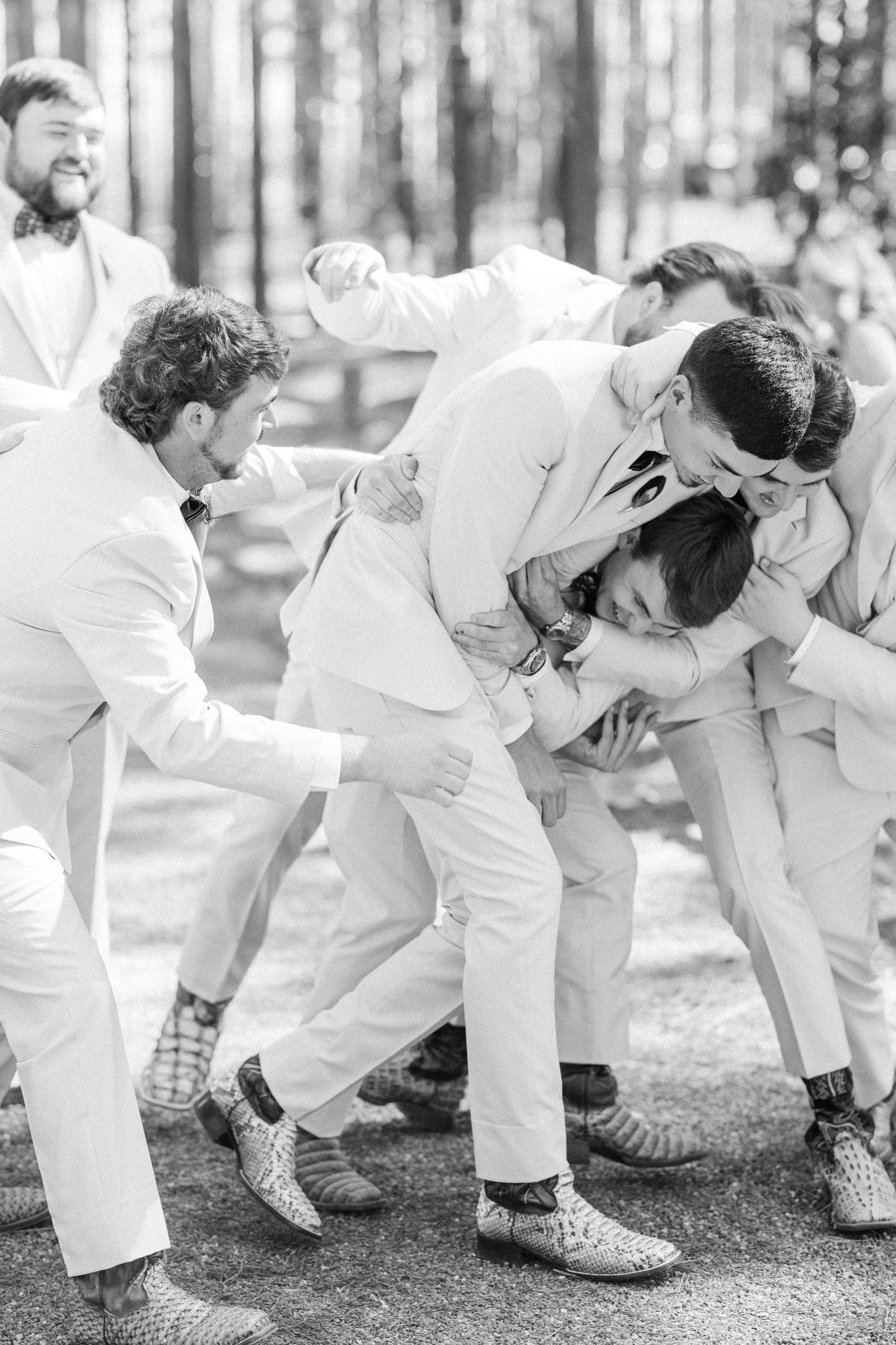 Groomsmen in white suits playfully wrestle outdoors at a woodland wedding in black and white photo.