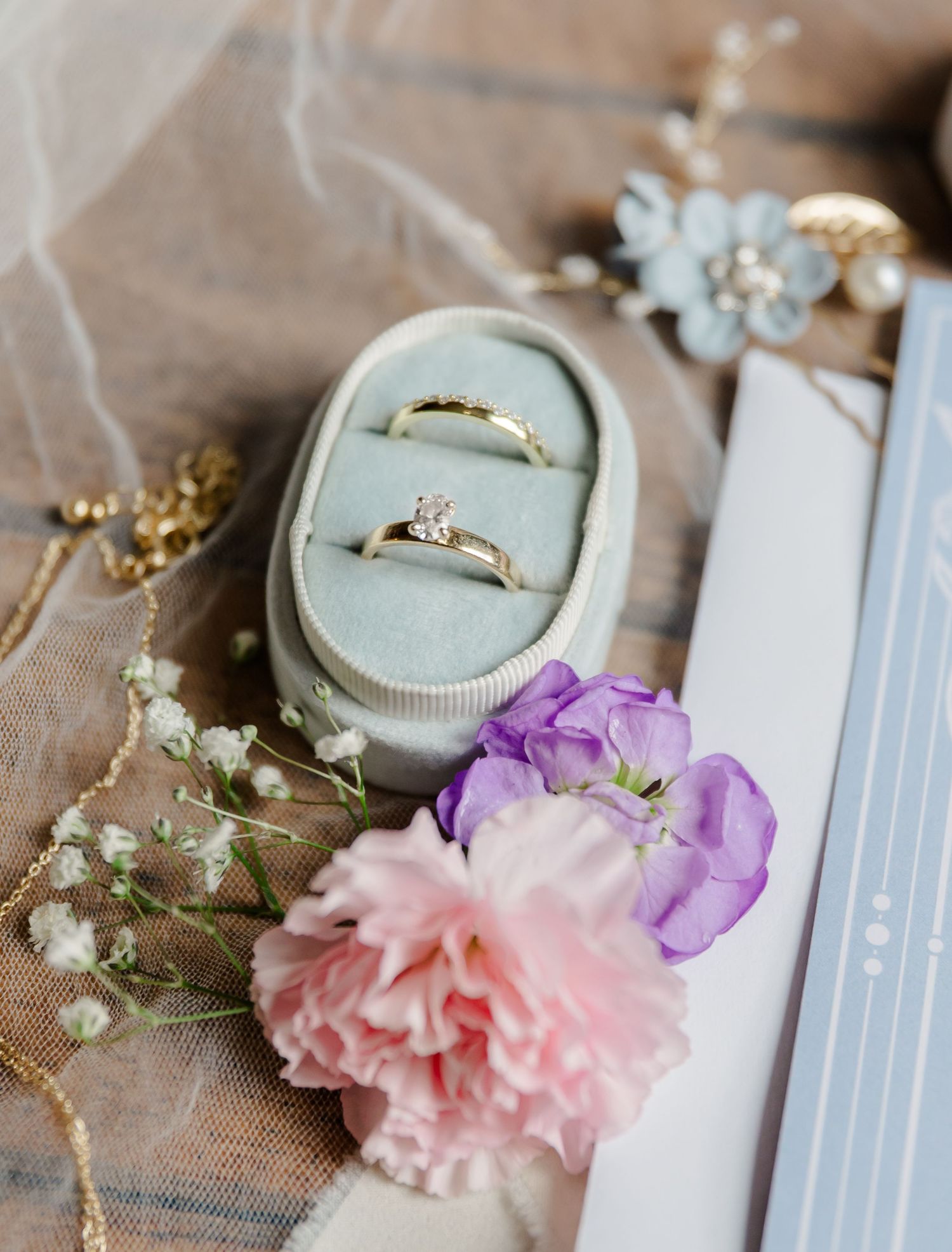 Wedding rings displayed in a light blue velvet ring box with pink carnation and purple flowers as decoration.