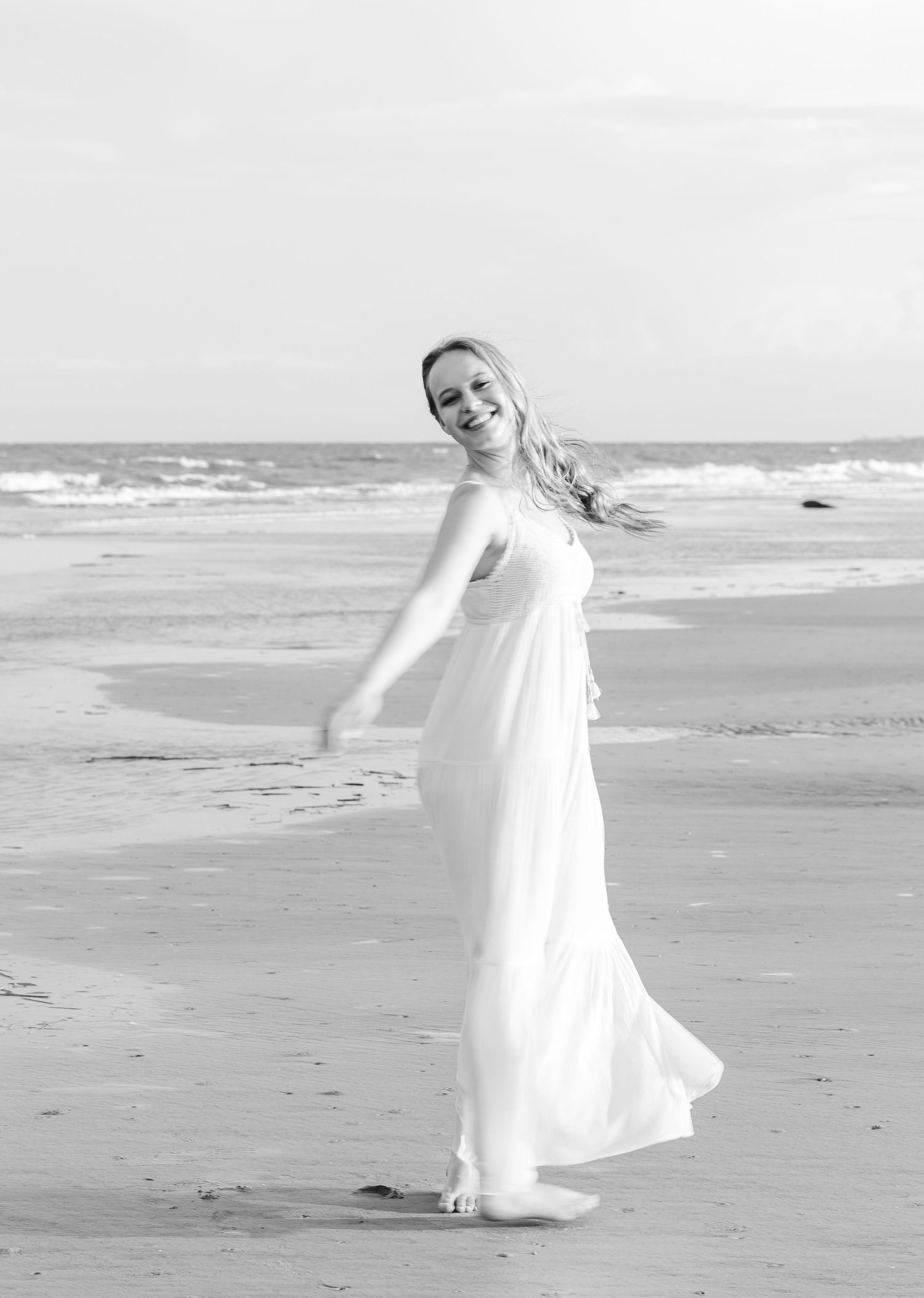 Ethereal black and white beach portrait with flowing white dress in ocean breeze.