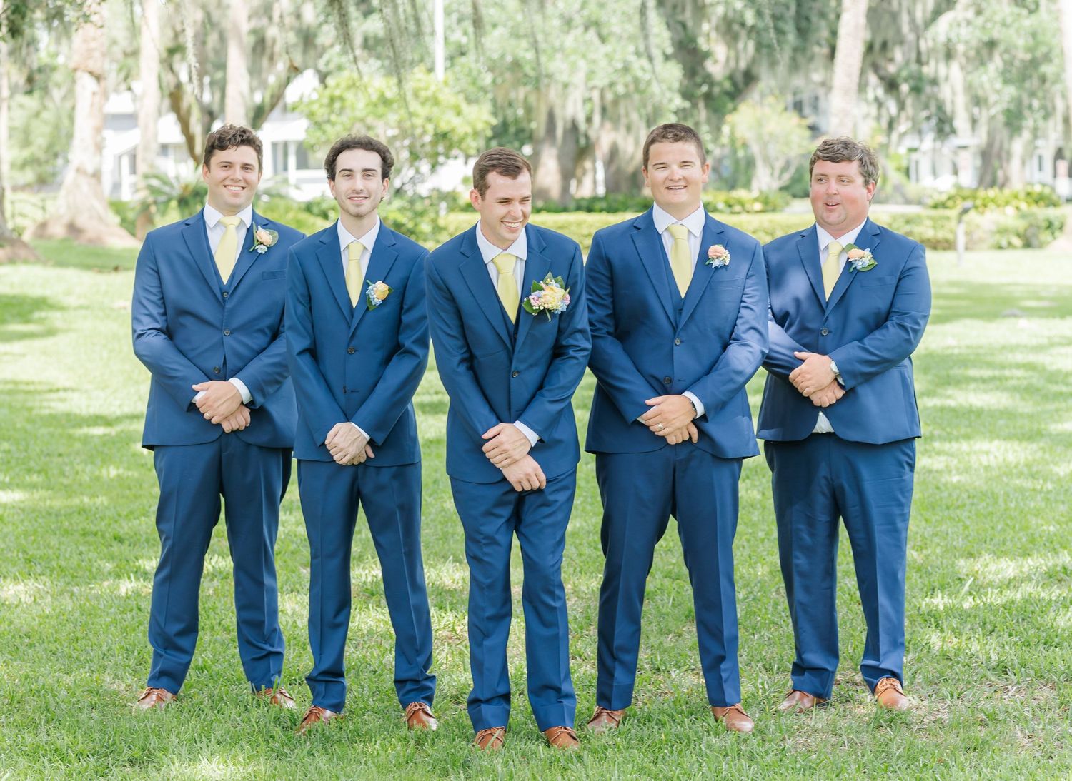 Groomsmen in matching navy blue suits stand together on a grassy lawn beneath Spanish moss trees.
