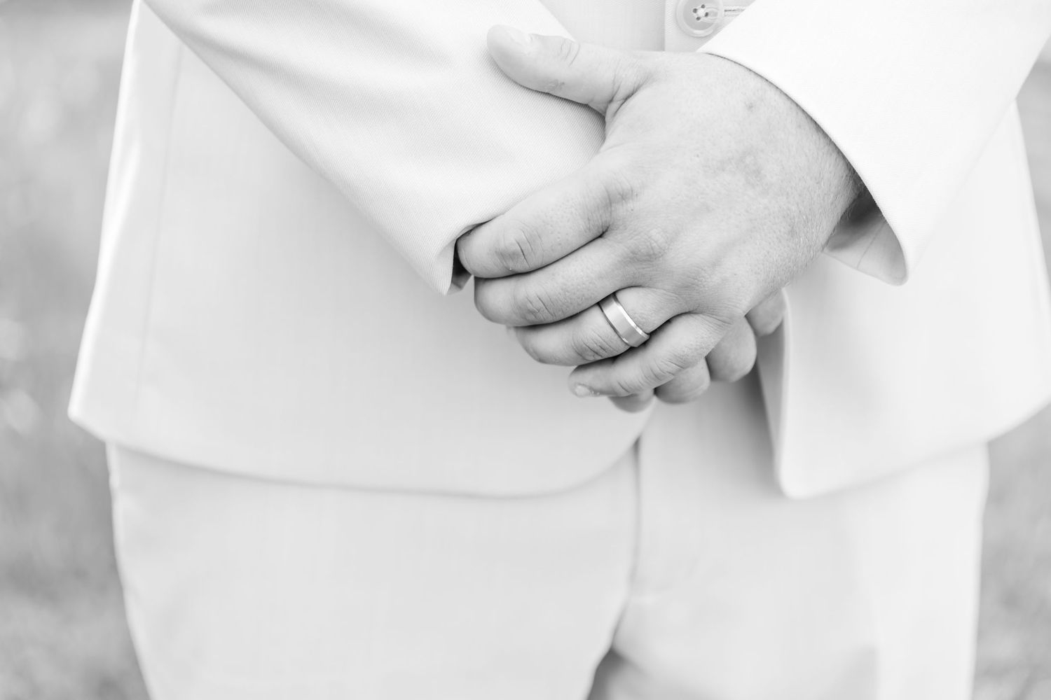 Black and white detail shot of groom's hands with wedding ring against white suit.