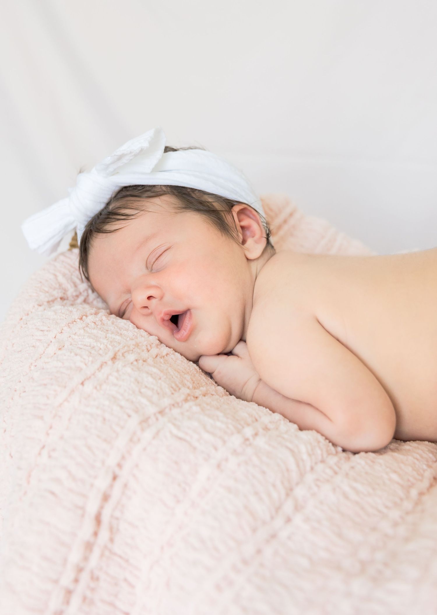 Sleeping newborn with white bow headband yawns while resting on pink blanket.