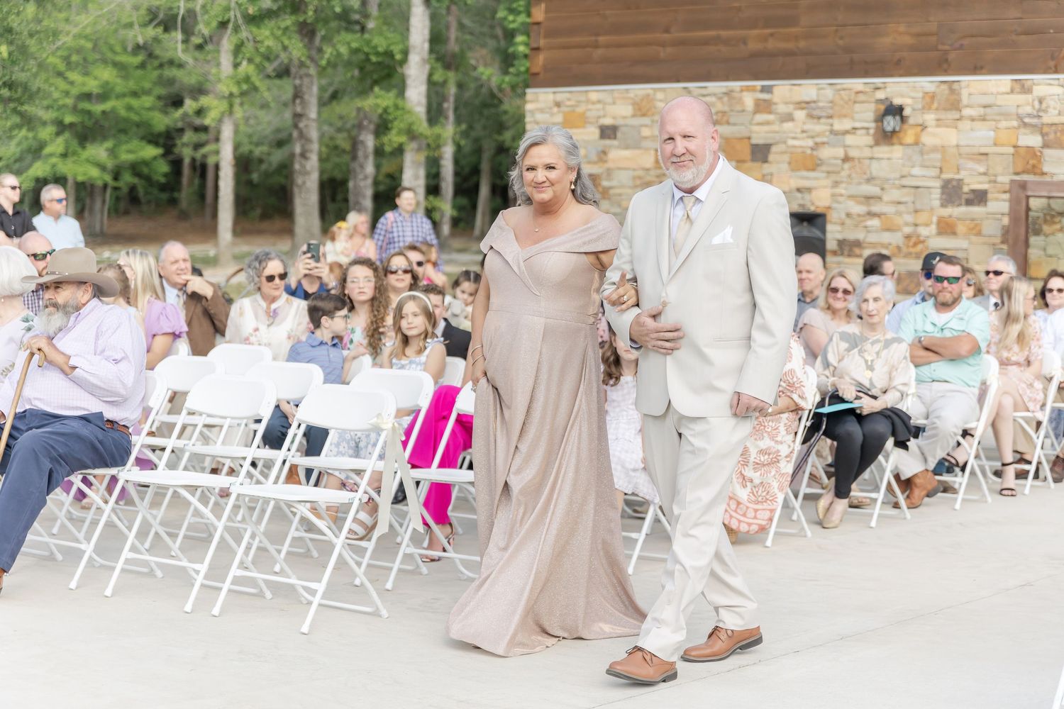 Wedding guests watch as a couple in neutral formal attire walk down an outdoor aisle at a rustic stone venue.