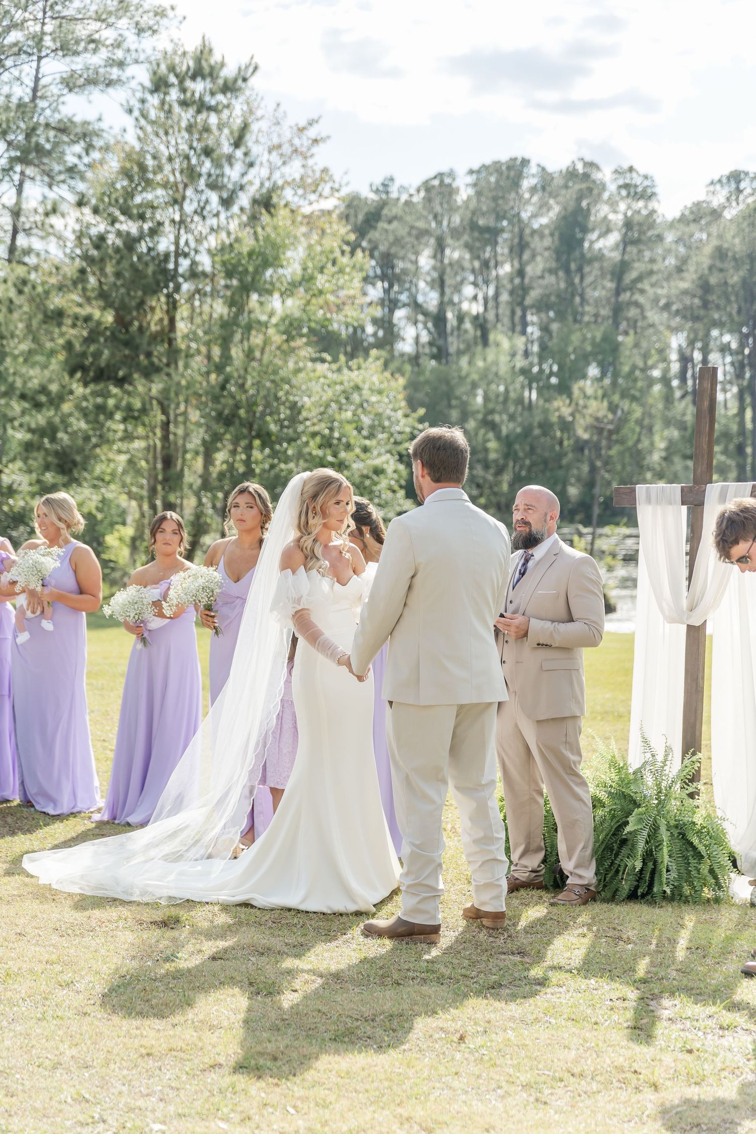 A sunlit outdoor wedding ceremony with white decorative columns and lush greenery arrangements.