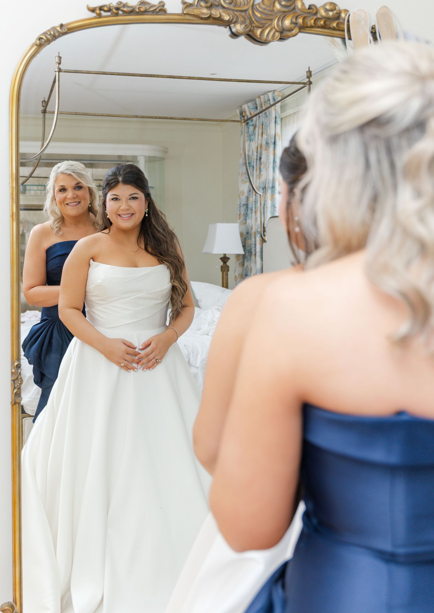 Bride in white strapless gown admires her reflection in an ornate gold mirror with bridesmaid in navy dress.