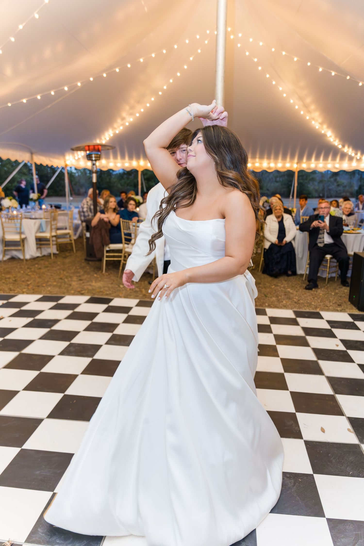 Bride in strapless white ball gown twirls on black and white checkered dance floor under lit wedding tent.