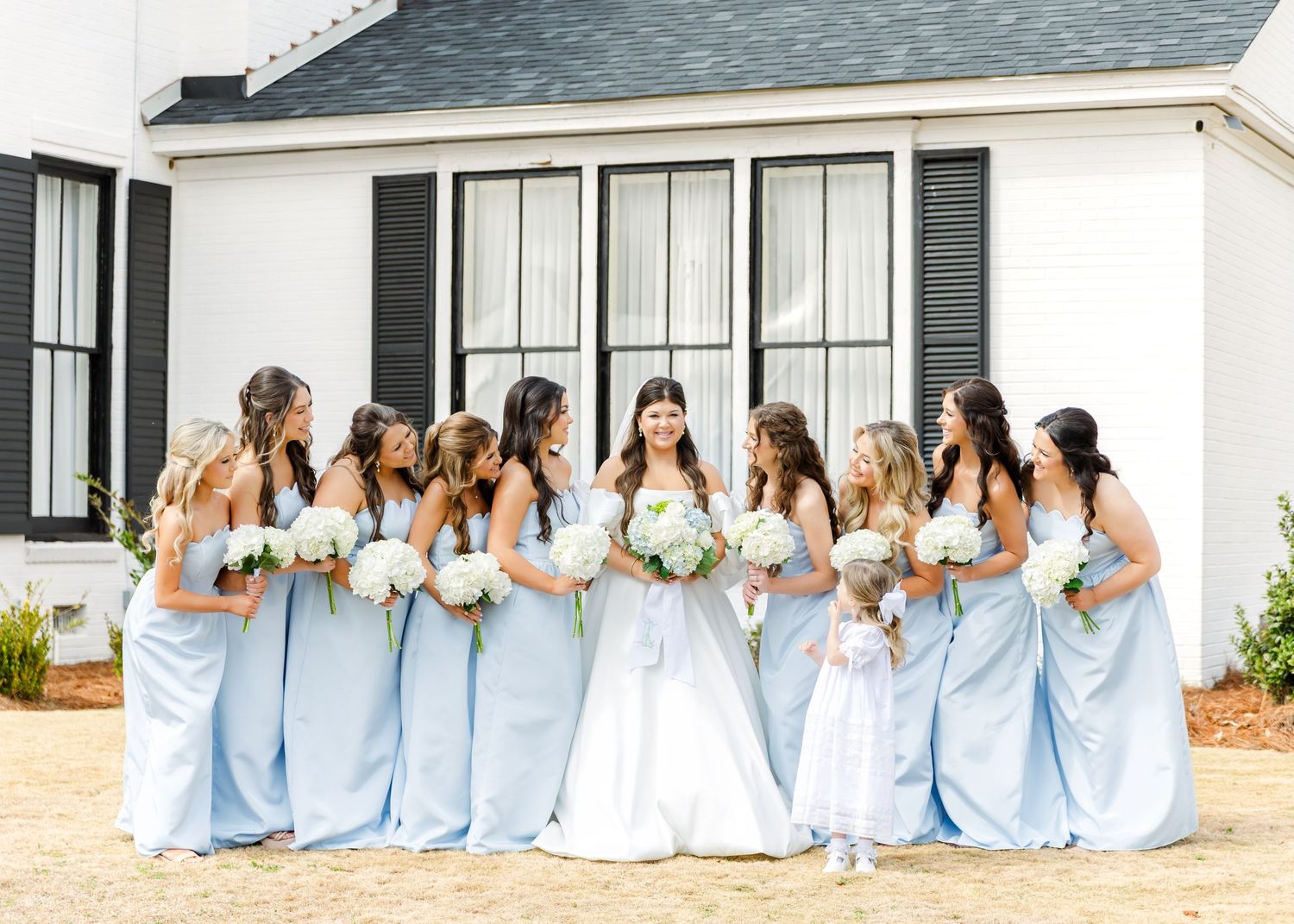 Bride surrounded by bridesmaids in light blue gowns holding white bouquets outside white wedding venue.