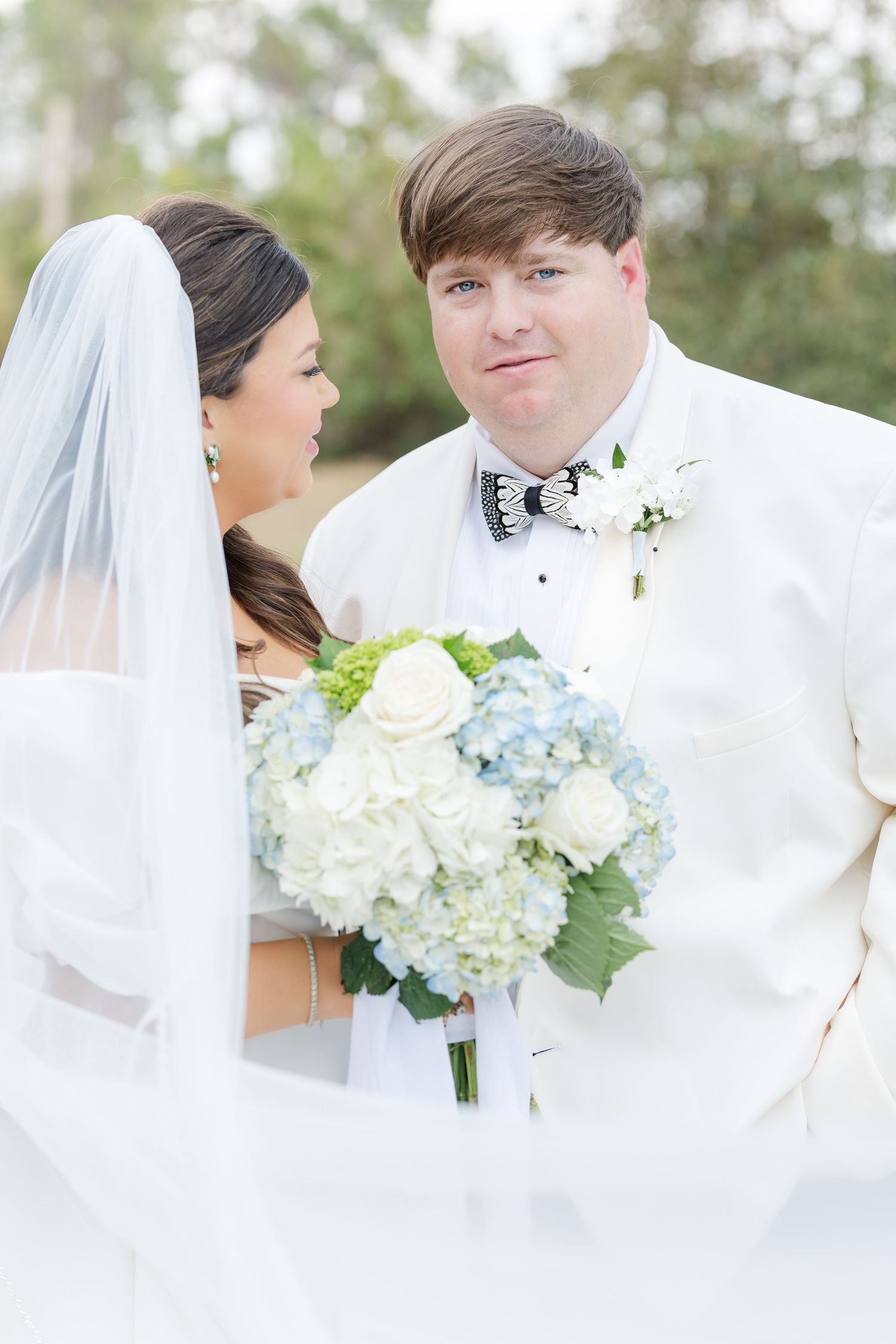 Bride and groom portrait with blue and white hydrangea bouquet, groom in white tuxedo with bow tie.
