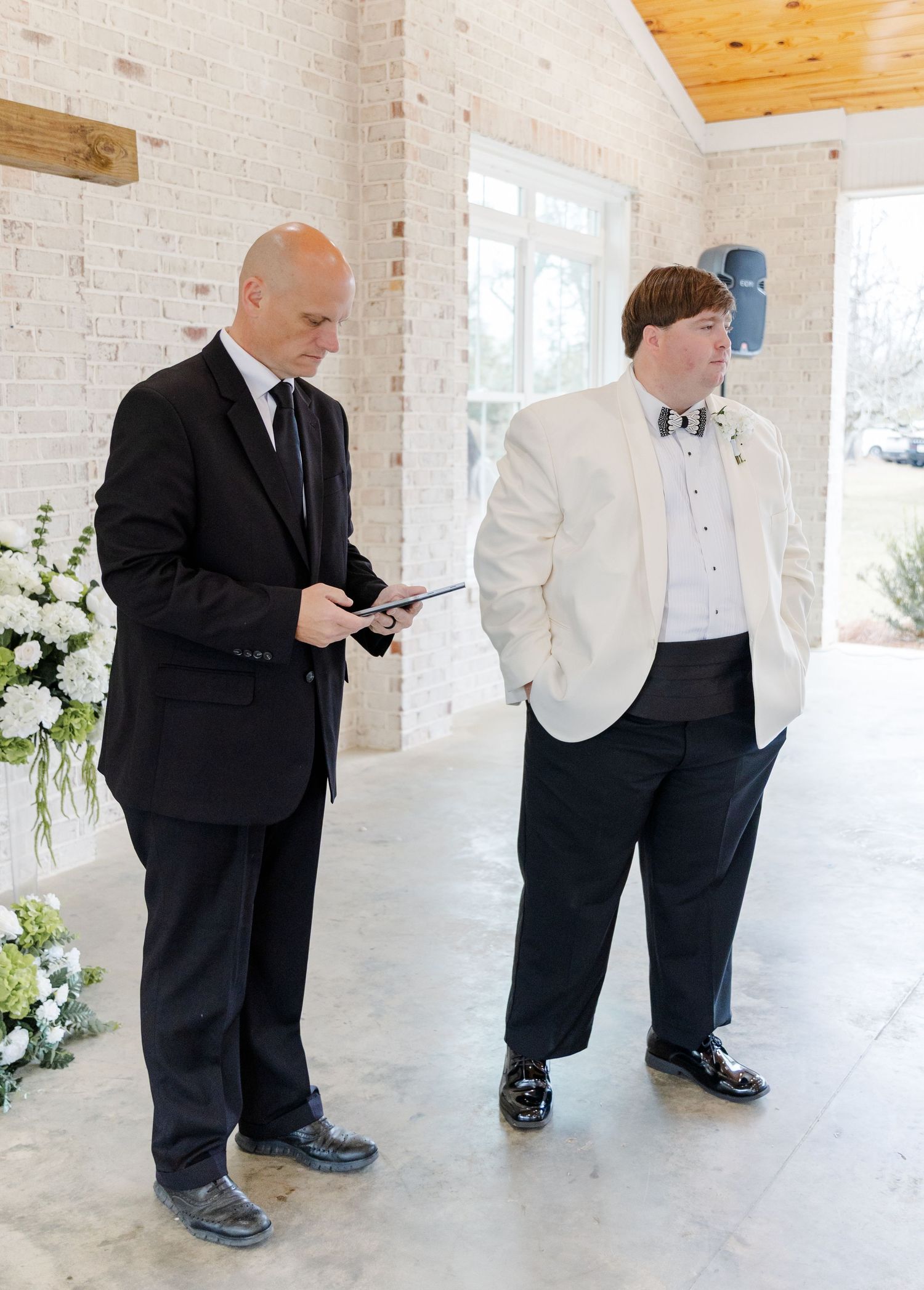 Groom in white tuxedo jacket stands nervously beside officiant holding tablet while awaiting bride at altar.