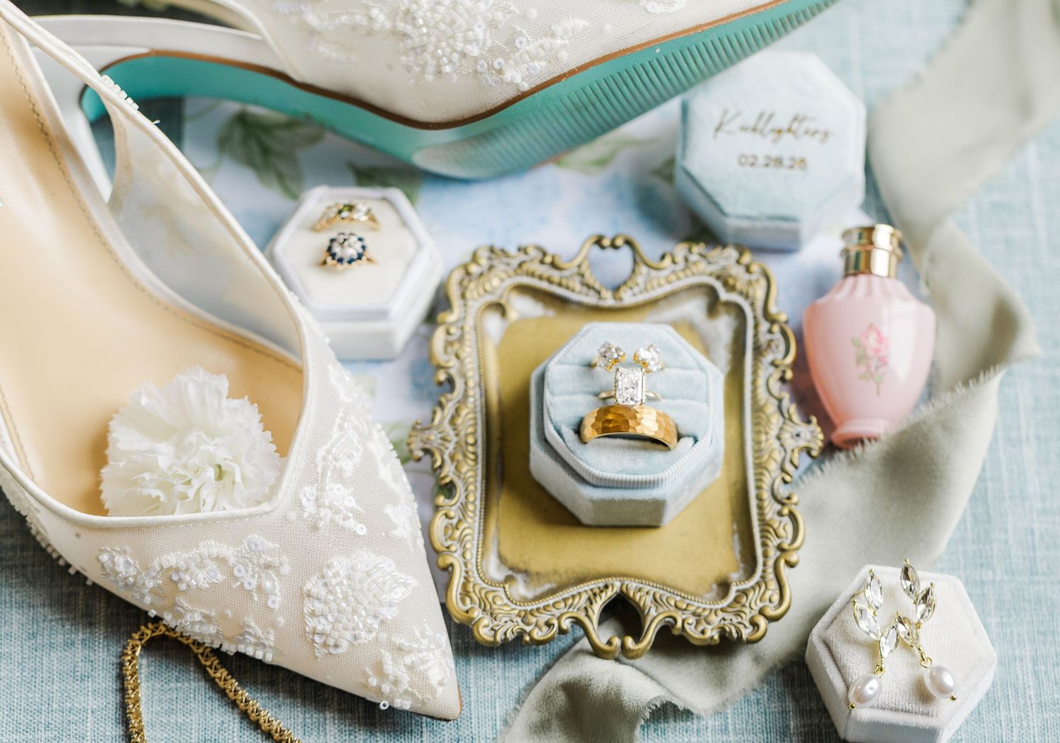 Elegant bridal flat lay with lace heels, gold wedding rings, velvet ring boxes, and pink perfume on blue backdrop.
