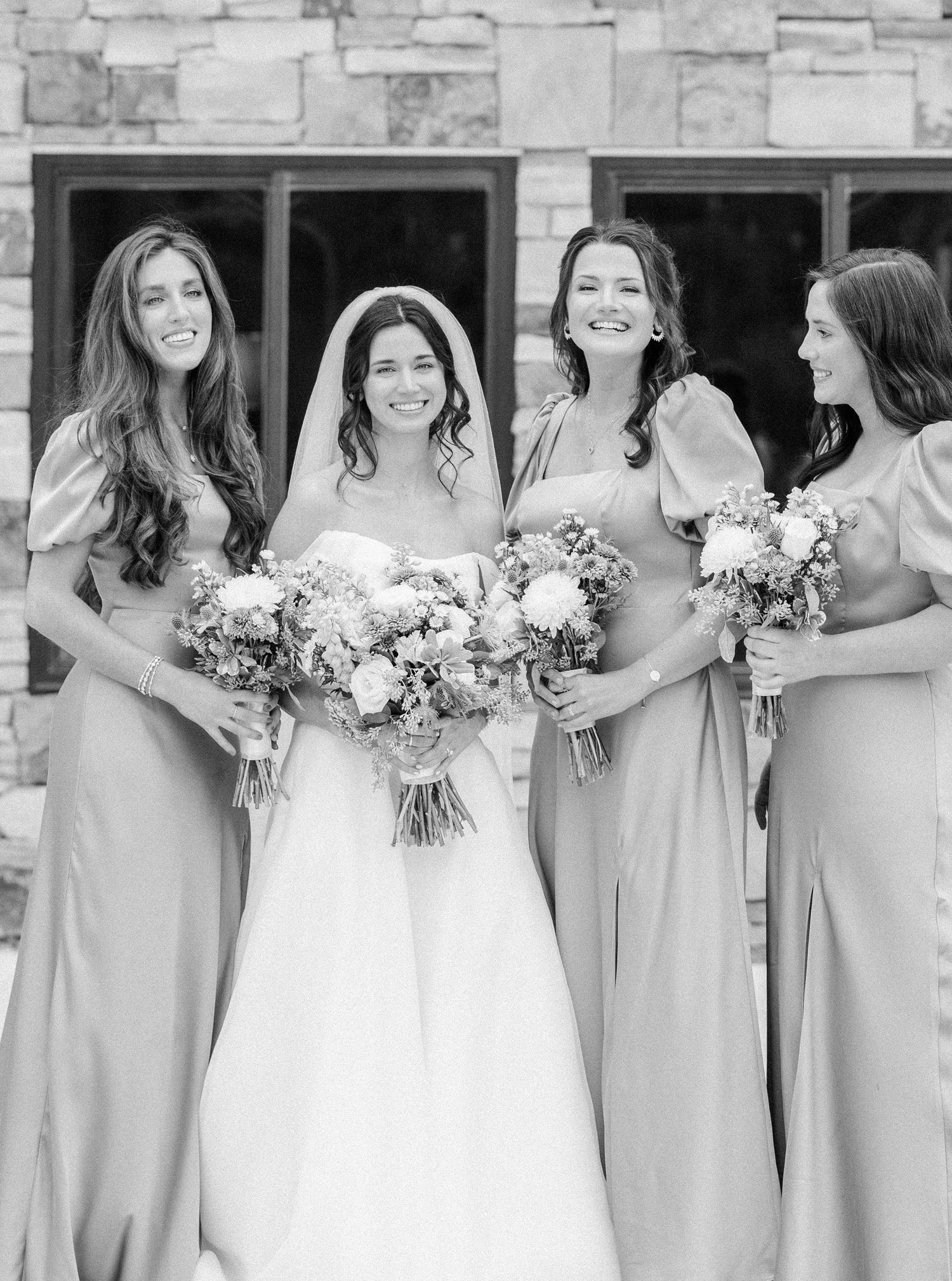 Black and white photo of bridesmaids in light colored dresses holding bouquets while standing with bride in white gown.