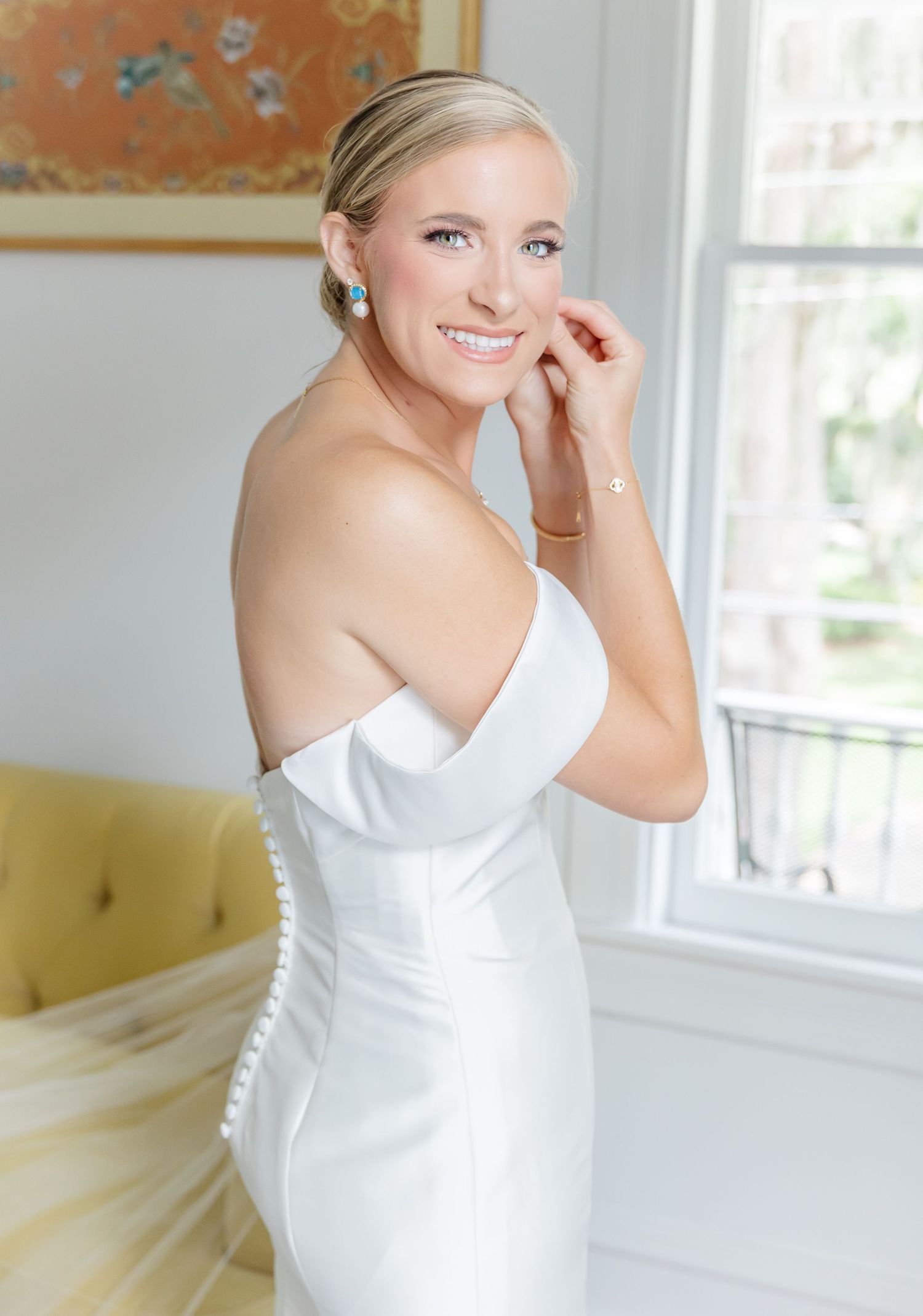 A bride in an elegant white strapless wedding gown with buttons down the back poses by a sunny window.