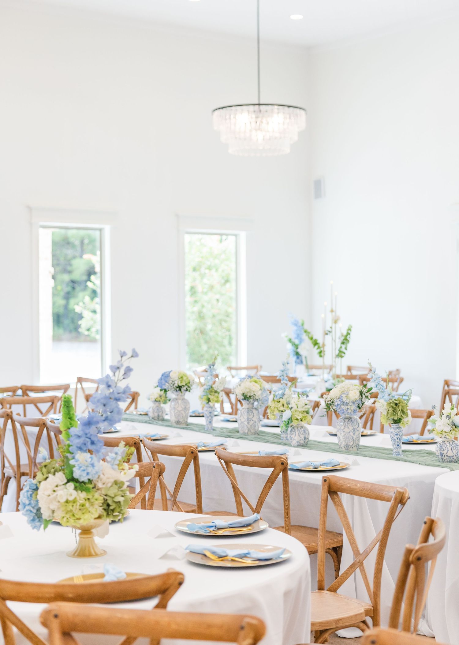 Elegant wedding reception with wooden cross-back chairs, white tablecloths, blue floral centerpieces, and a crystal chandelier.