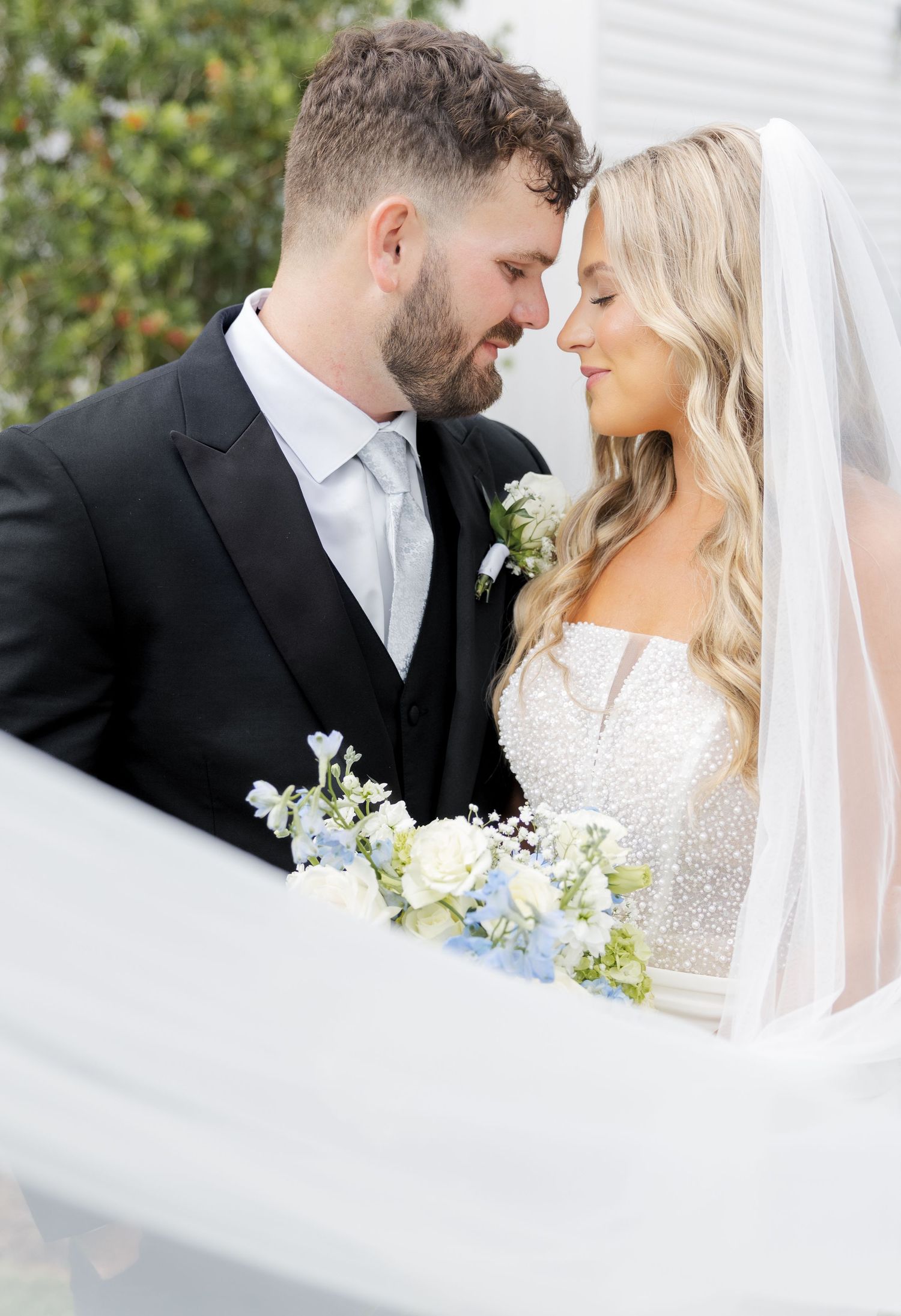 Bride and groom share an intimate moment under a veil, holding blue and white wedding bouquet.