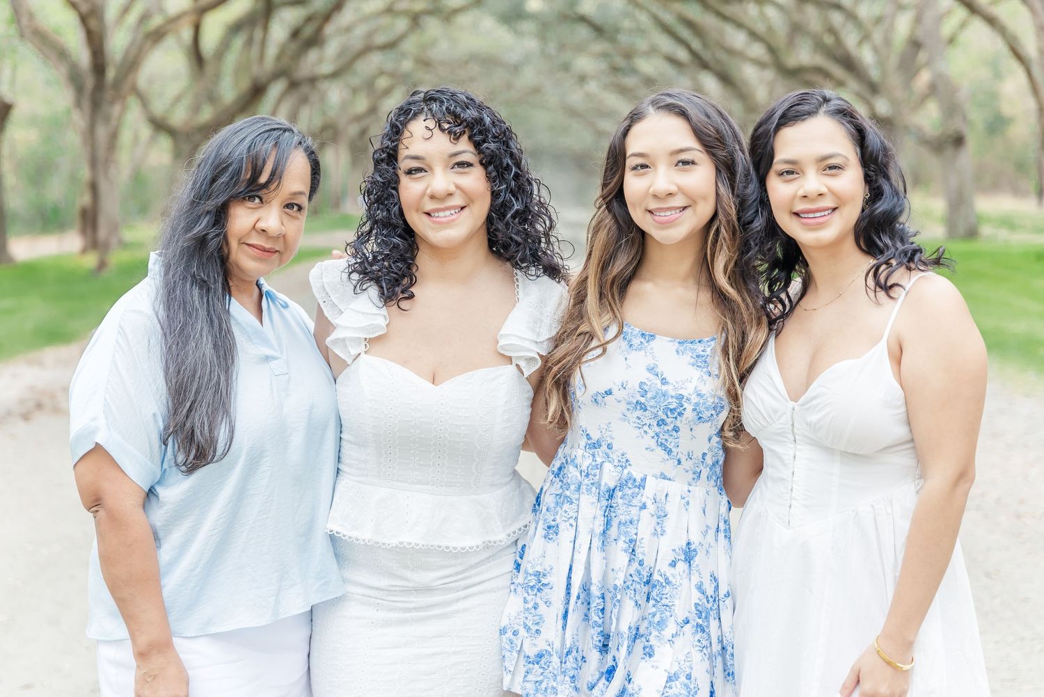 Four women smile together in coordinating white and blue outfits under oak trees.