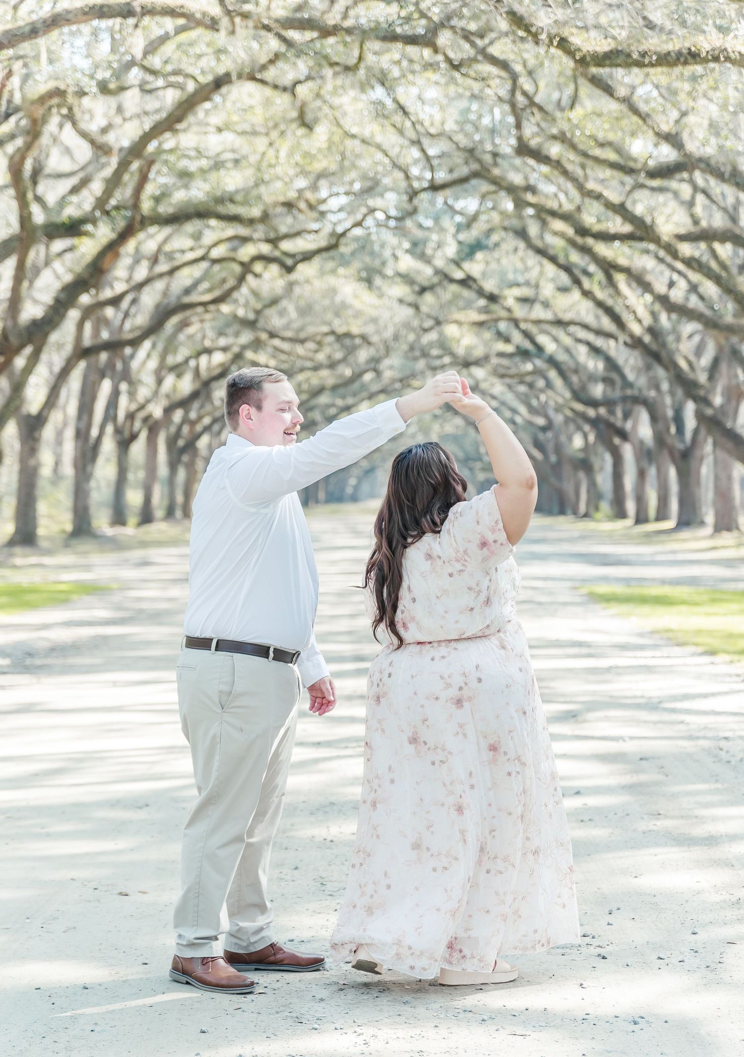 A couple dancing together on a sunlit path beneath arching oak trees.