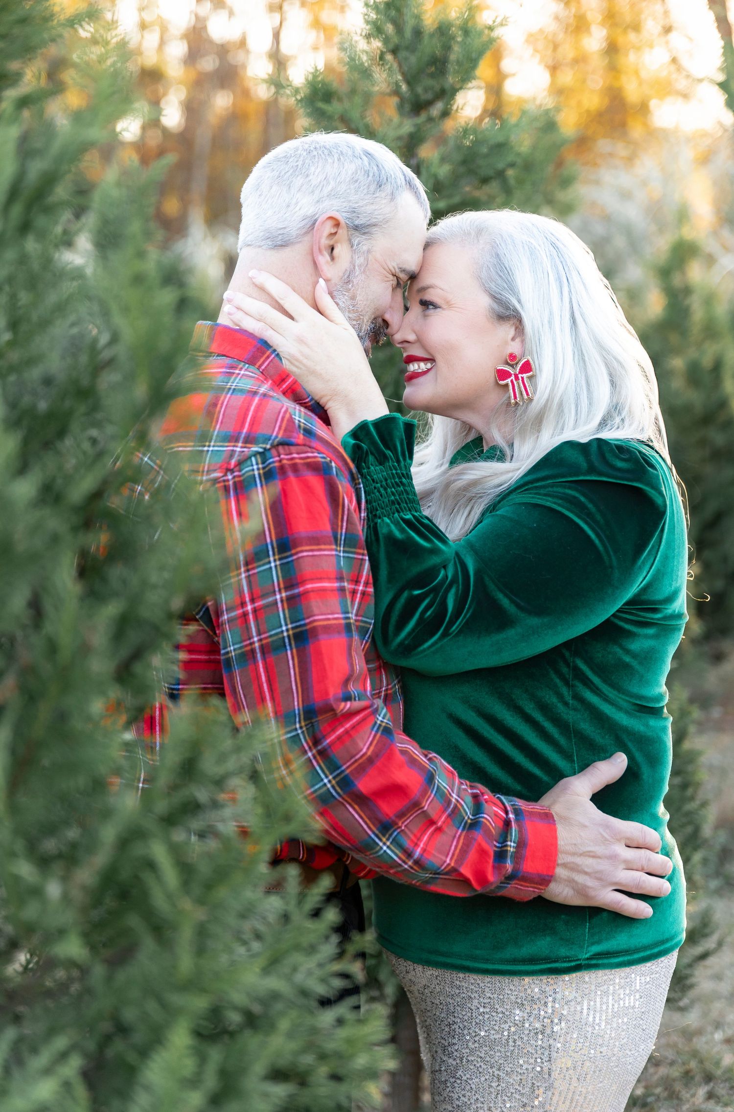 Intimate moment between couple in Christmas-themed outfits embracing among evergreen trees.