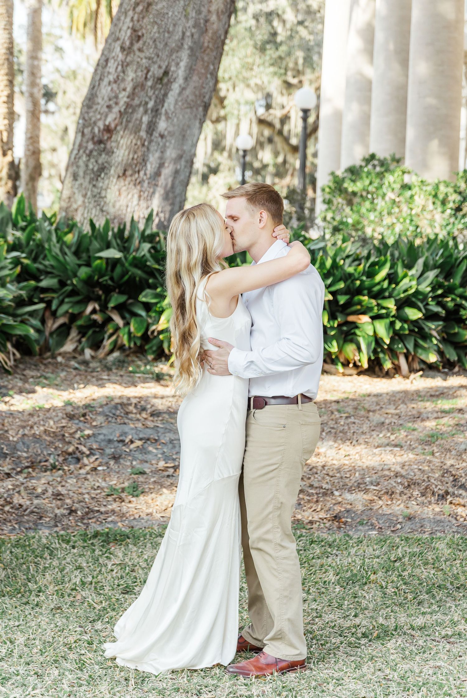 Couple embraces in white wedding attire against backdrop of columns and palm trees.