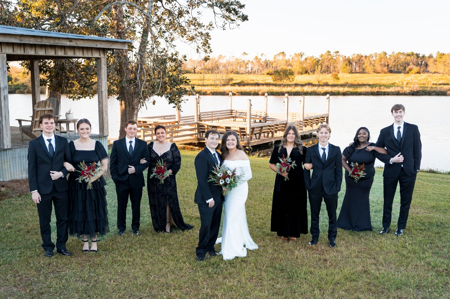 Full wedding party poses outdoors with lake and dock in background at golden hour.