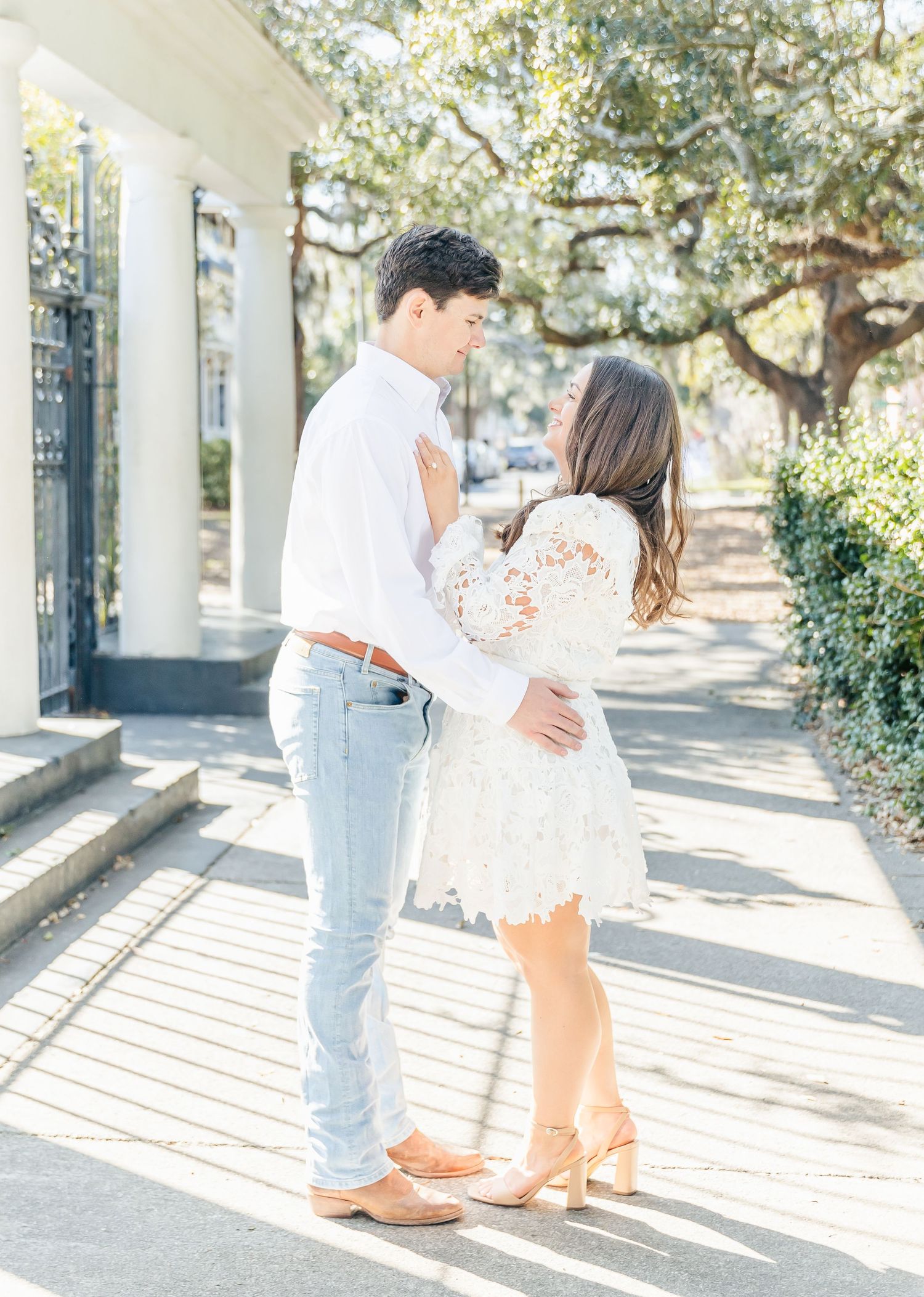 Romantic couple embraces in sunlit garden pathway wearing white and denim casual attire.