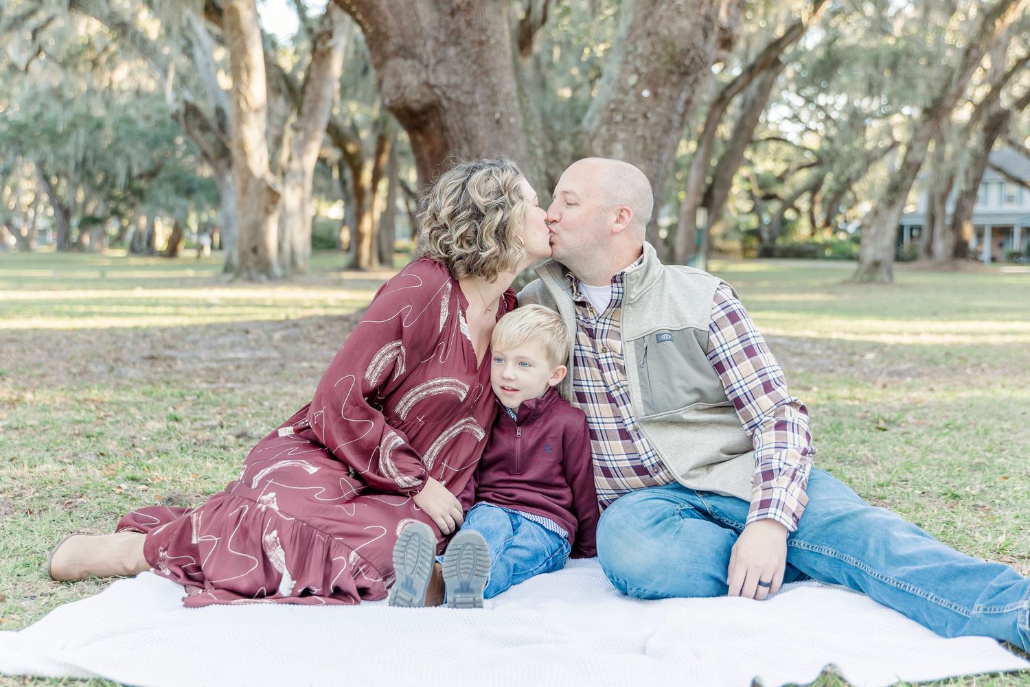 A family shares an affectionate moment while sitting on a white blanket in a park.