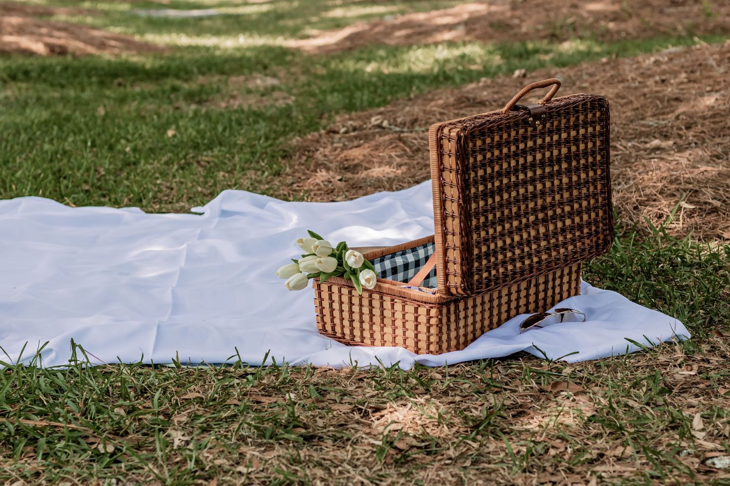 Vintage wicker picnic basket opened on a white blanket in a park setting.
