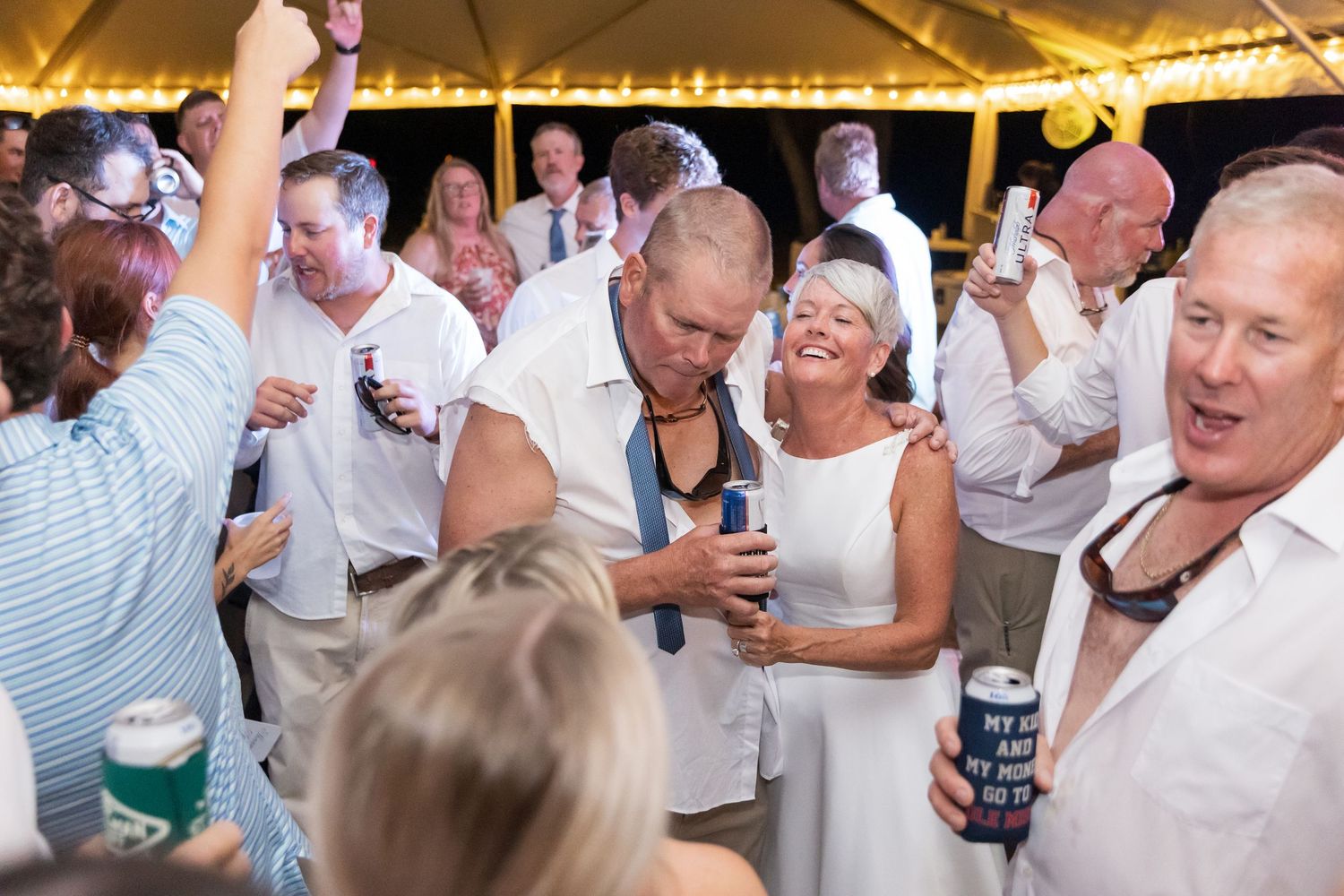 Festive party crowd dancing and mingling under yellow tent lighting at evening celebration.