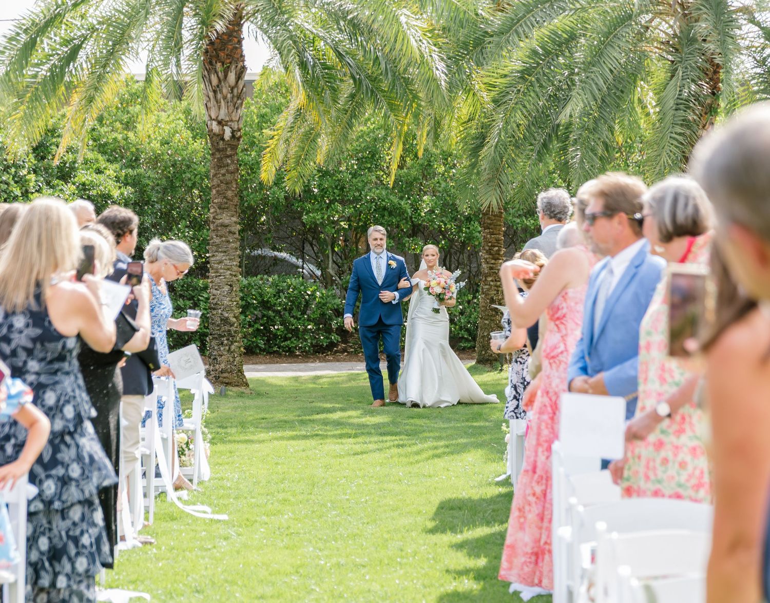 Wedding guests in floral dresses gather on a lush green lawn surrounded by tropical palm trees during an outdoor ceremony.