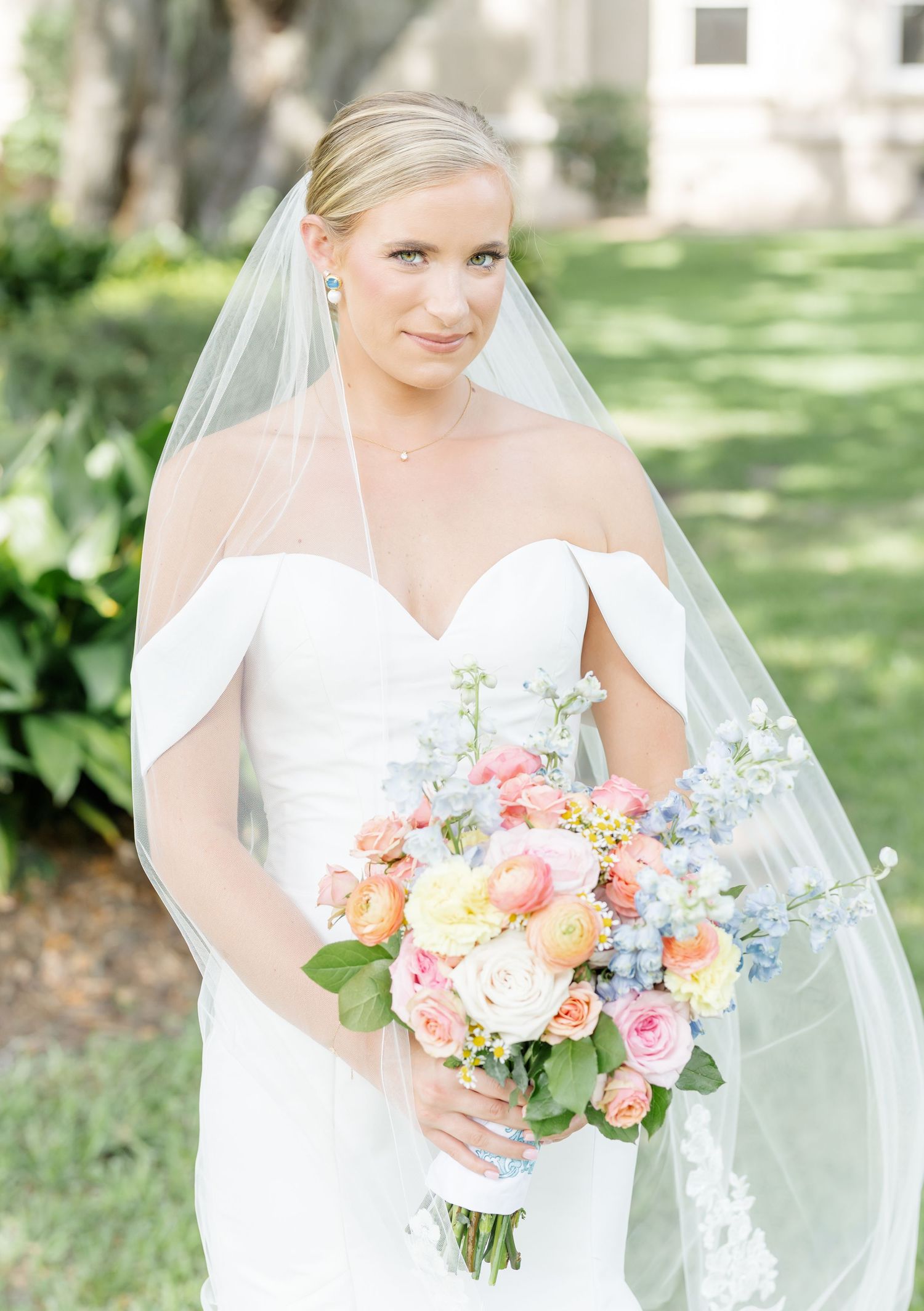 Elegant bride in off-shoulder white gown holds stunning pastel bouquet with roses and delicate flowers in garden setting.