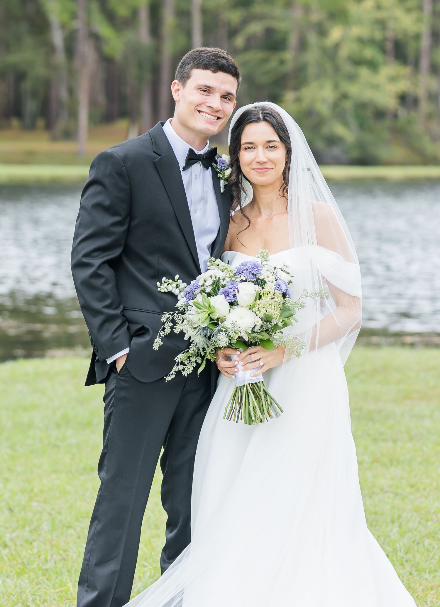 A bride and groom pose together by a lake, with the bride holding a beautiful blue and white floral bouquet.
