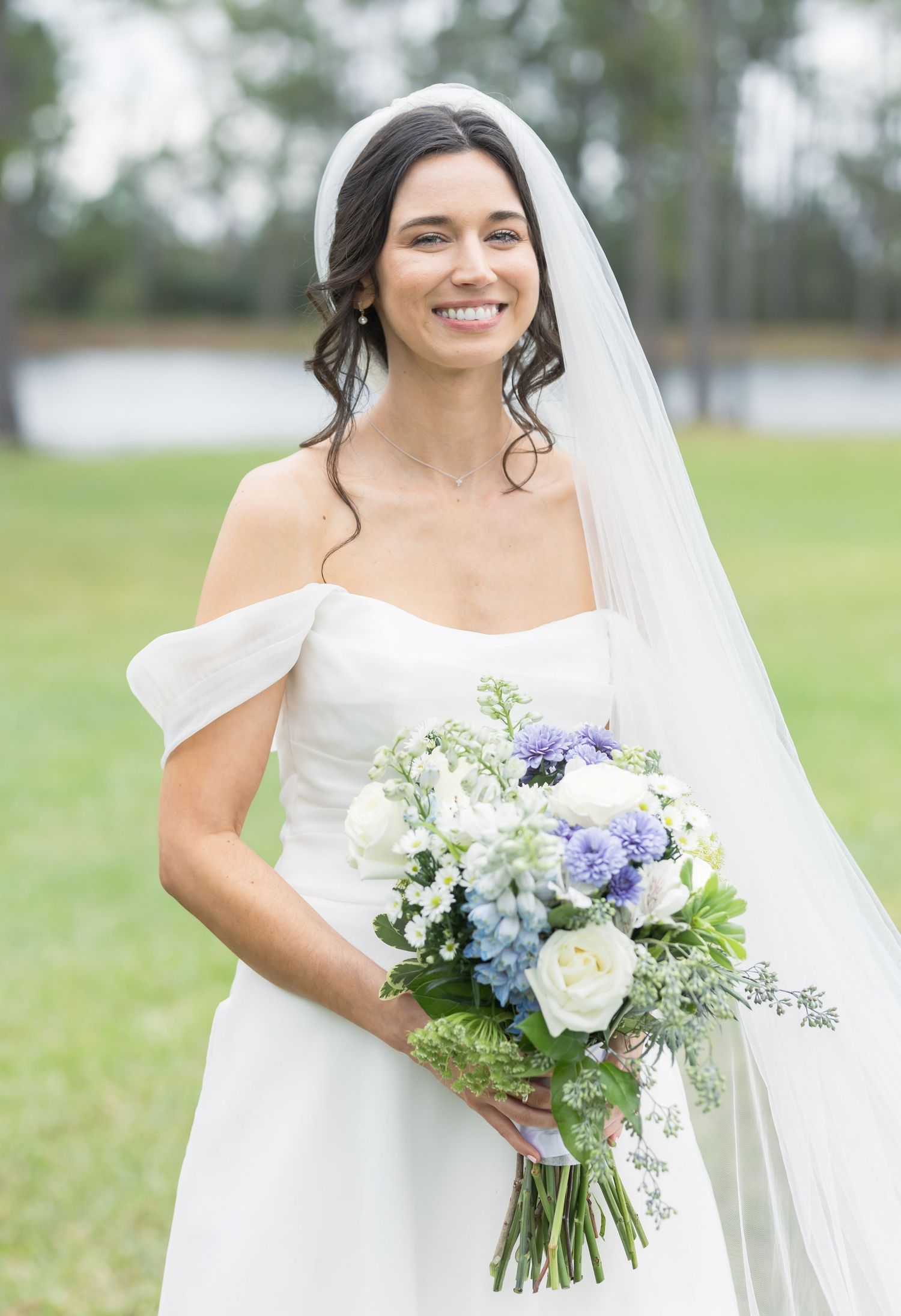 Bride in elegant white wedding dress holds stunning bouquet of white and purple flowers against a pastoral outdoor setting.