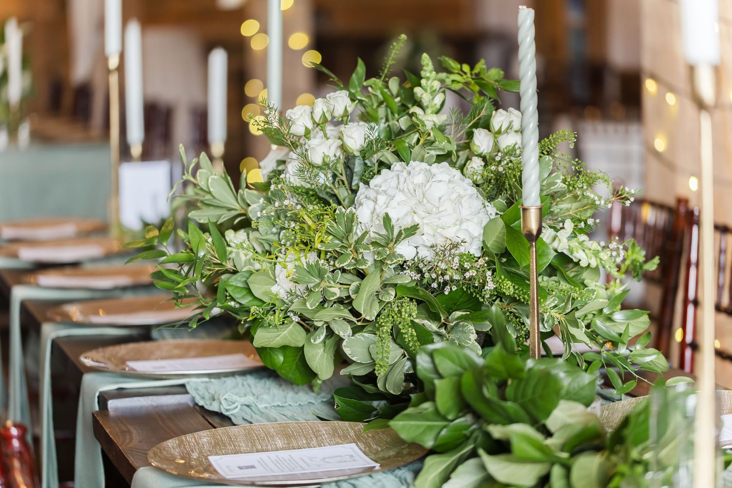 Elegant wedding reception table setting with white floral centerpieces and greenery arrangements.