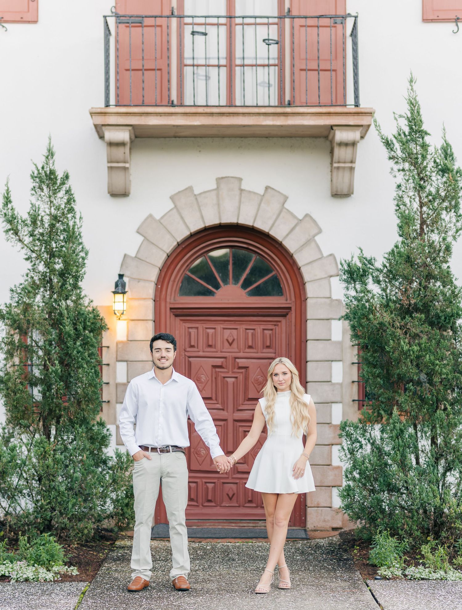 A couple in light-colored outfits poses against a white building with red doors and green plants.