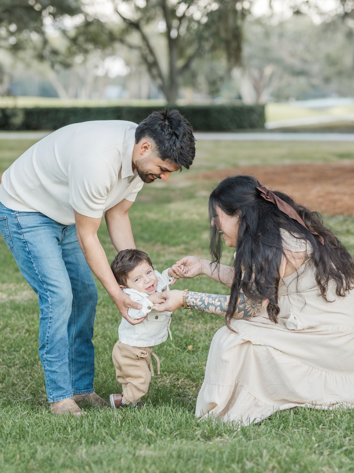 Family plays together on grass with their toddler learning to walk between parents.