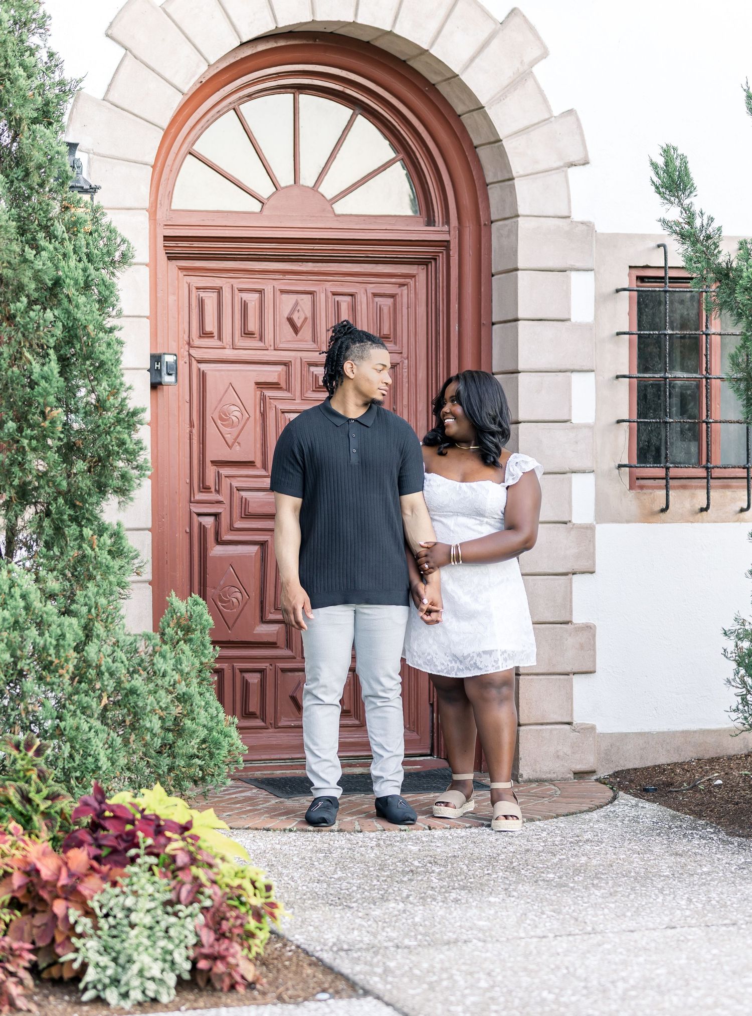 Couple standing together before an ornate red arched door surrounded by manicured shrubs.