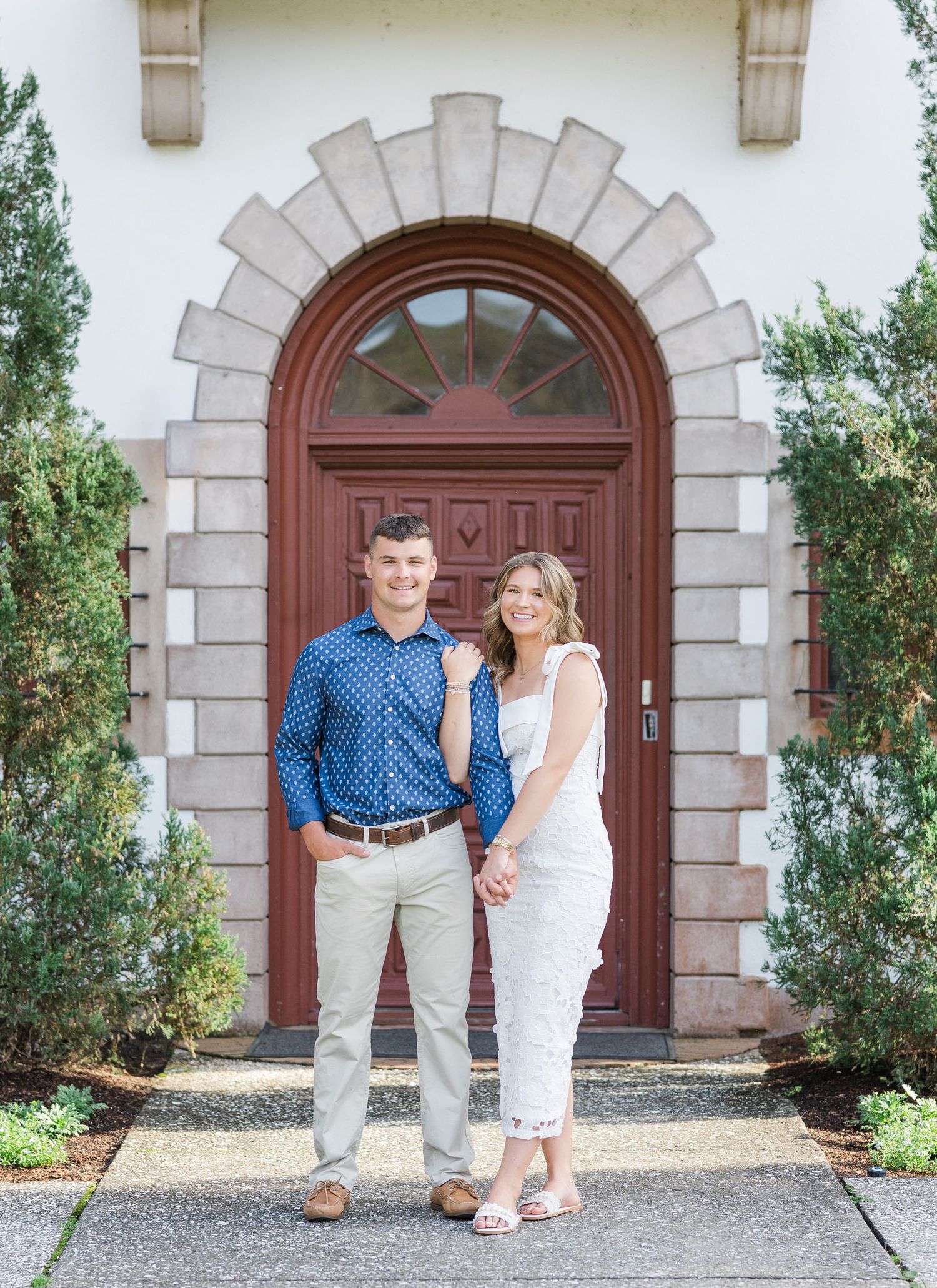 Couple holding hands smiling in front of elegant arched red door with stone accents and lush greenery.