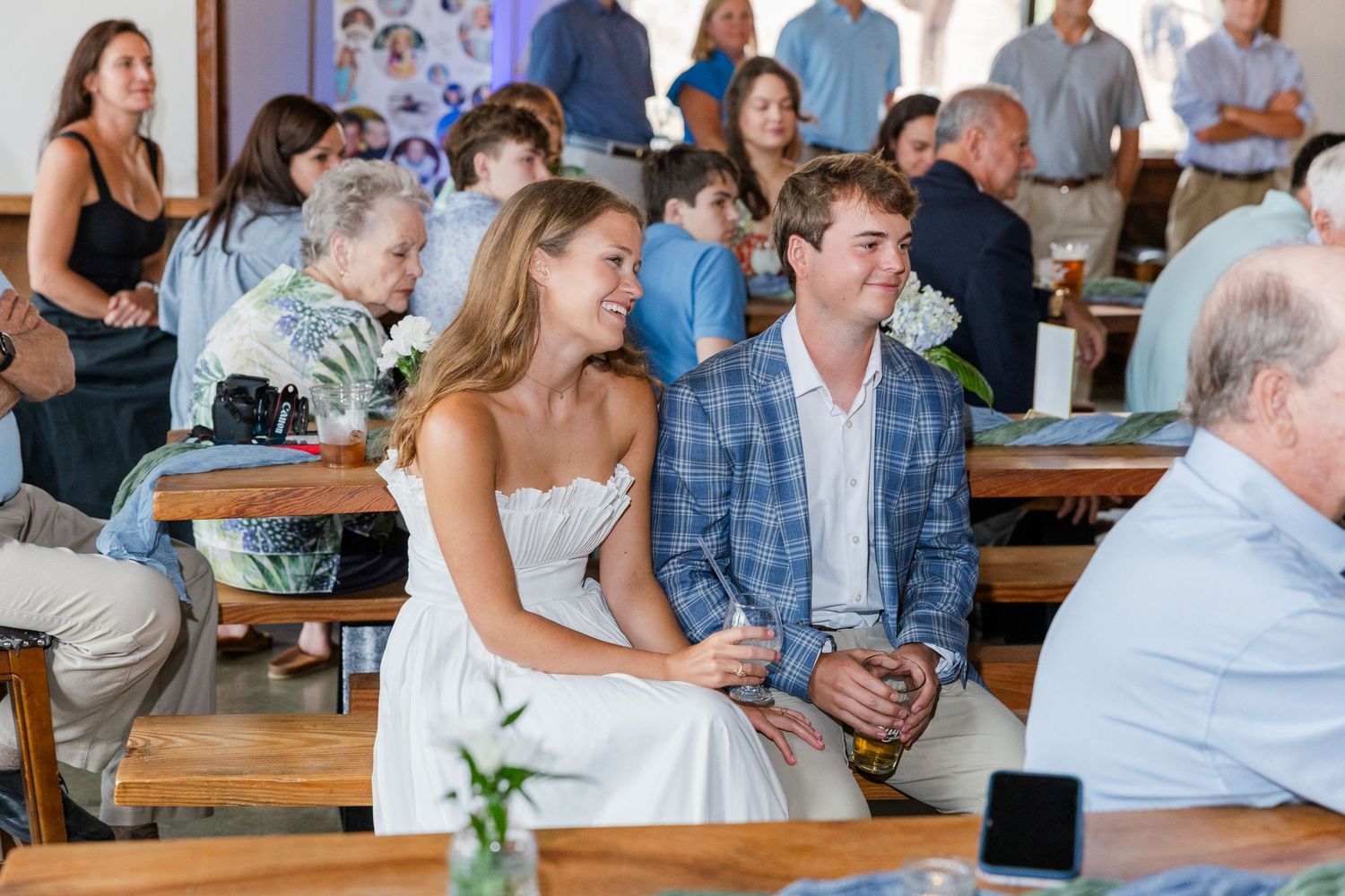 A bride in a white strapless dress and groom in a blue plaid suit smile joyfully at an indoor wedding reception.
