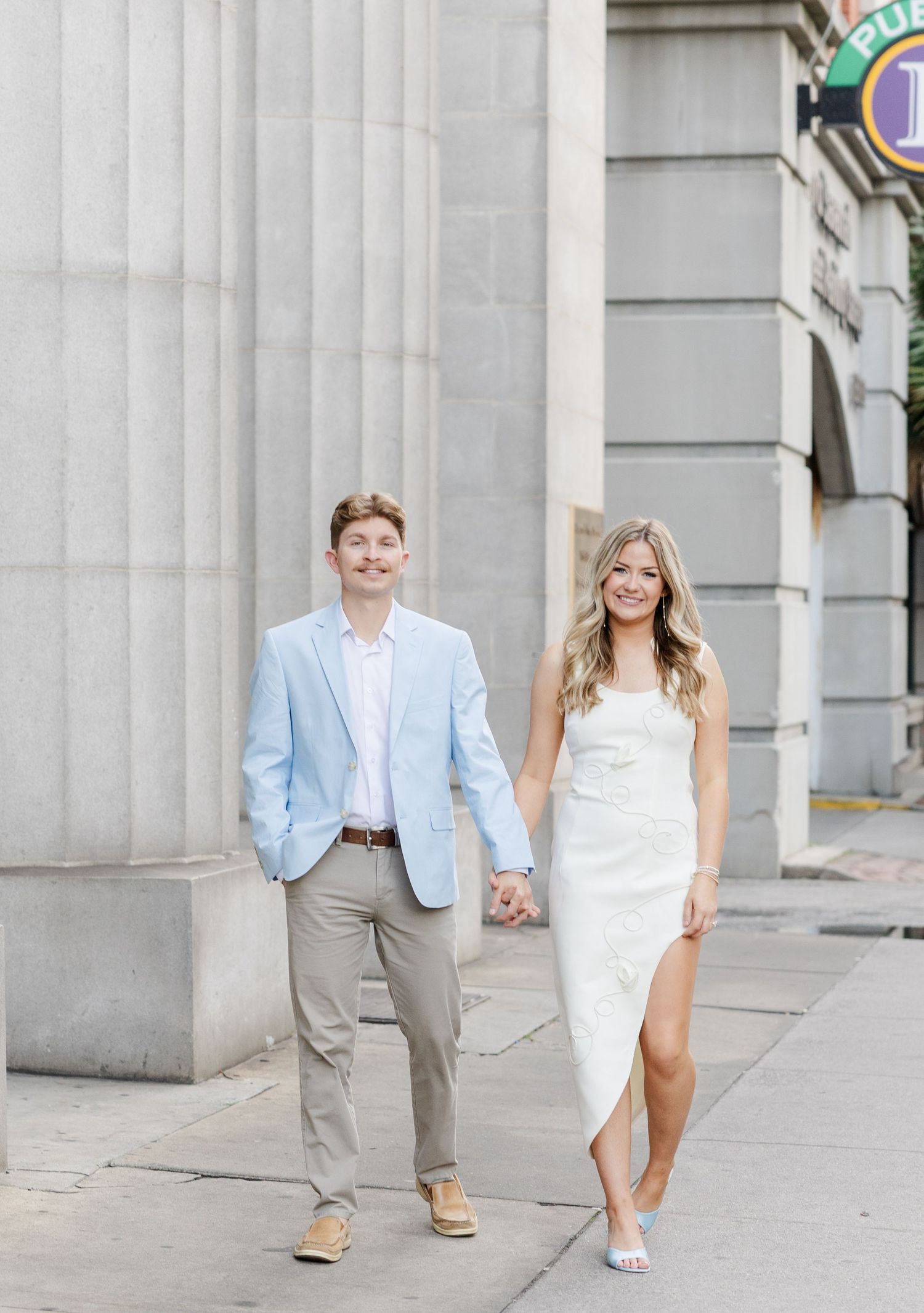Couple holding hands walking on urban sidewalk, man in light blue suit, woman in white slit dress.