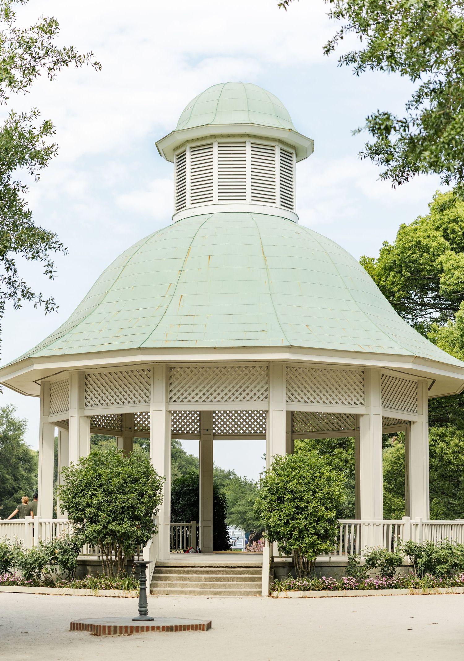 A white octagonal gazebo with a green copper dome roof surrounded by lush trees in a park setting.