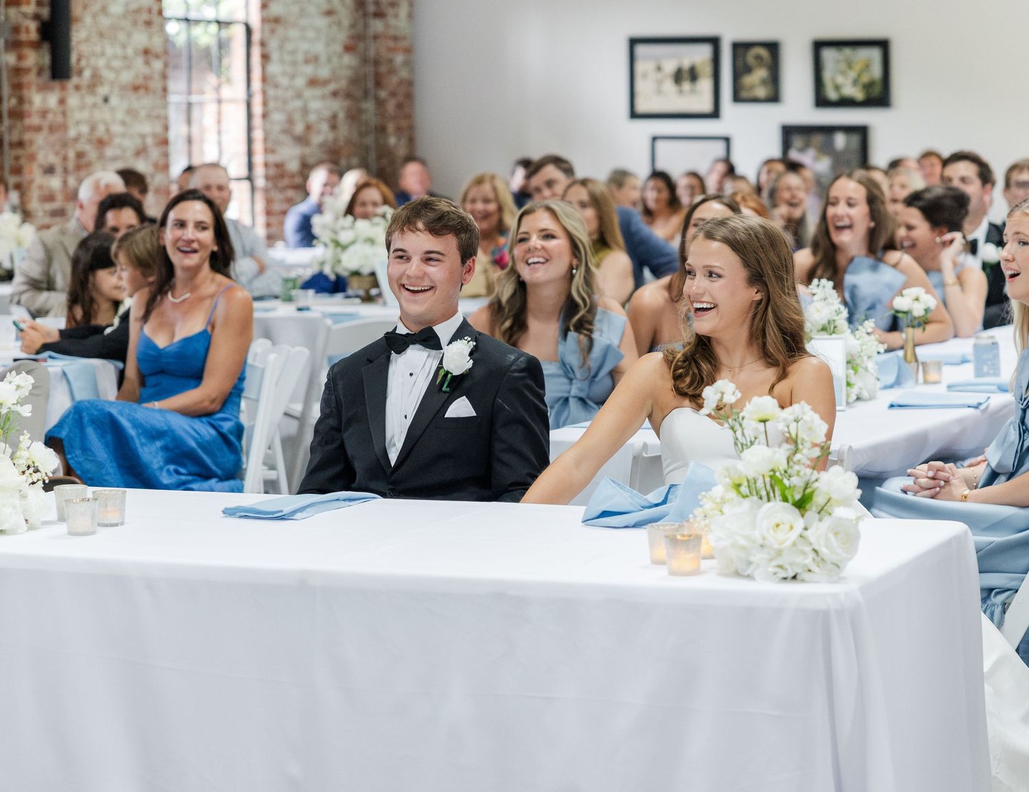 Bride and groom laugh at sweetheart table surrounded by guests at elegant wedding reception with white floral centerpieces.