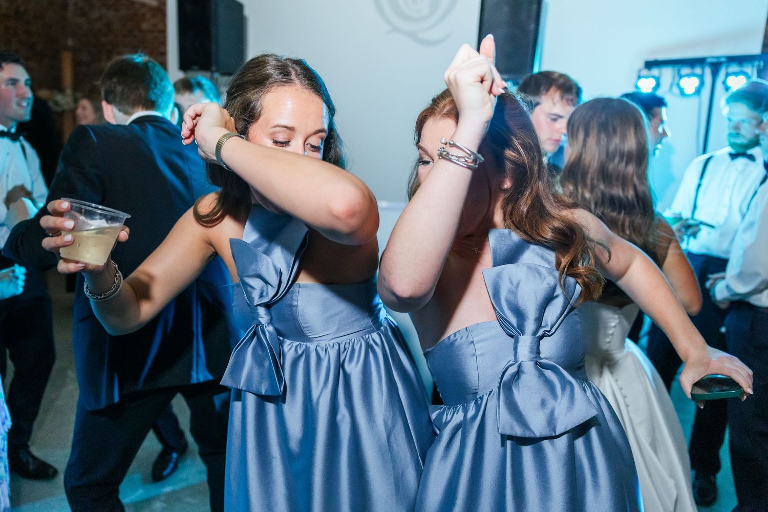 Two bridesmaids in matching blue dresses with bows dance together at wedding reception.